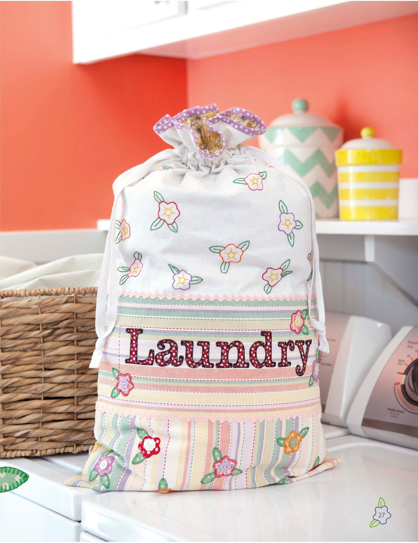 A vibrant drawstring laundry bag from the Salt Water Taffy Collection by Leisure Arts, featuring floral patterns and the word “Laundry” inspired by heirloom quilts and delicate hand embroidery, sits in a cheerful laundry room alongside a basket, jar, and striped container in the background.