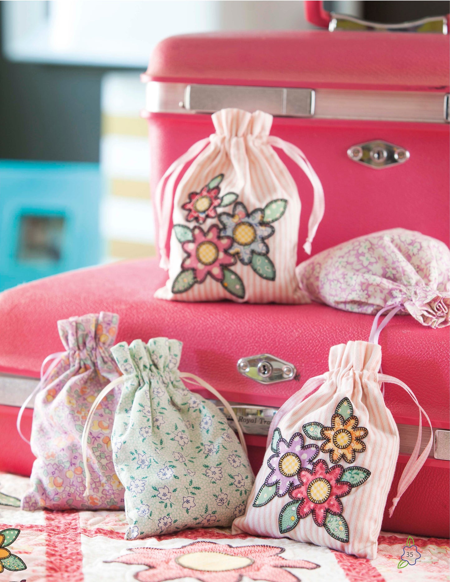 Four small fabric drawstring bags from the Salt Water Taffy Collection by Leisure Arts—two featuring floral appliqué and delicate hand embroidery—are arranged on and next to a pink suitcase. These pastel-colored bags, showcasing charming floral patterns reminiscent of heirloom quilts, rest with a partially visible quilt beneath them.