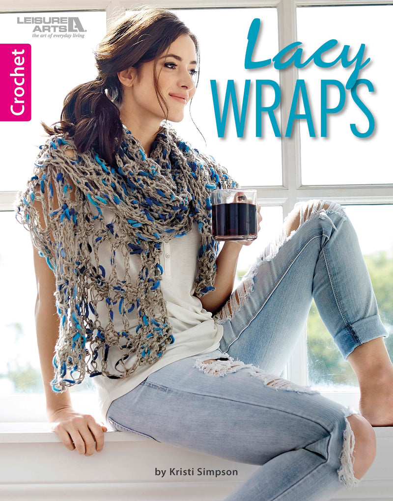 A woman in a blue and beige crocheted wrap from Lacy Wraps by Leisure Arts sits on a windowsill with a mug. She has long dark hair, a white shirt, and ripped light blue jeans. The book features chic lacy crochet shawl patterns.