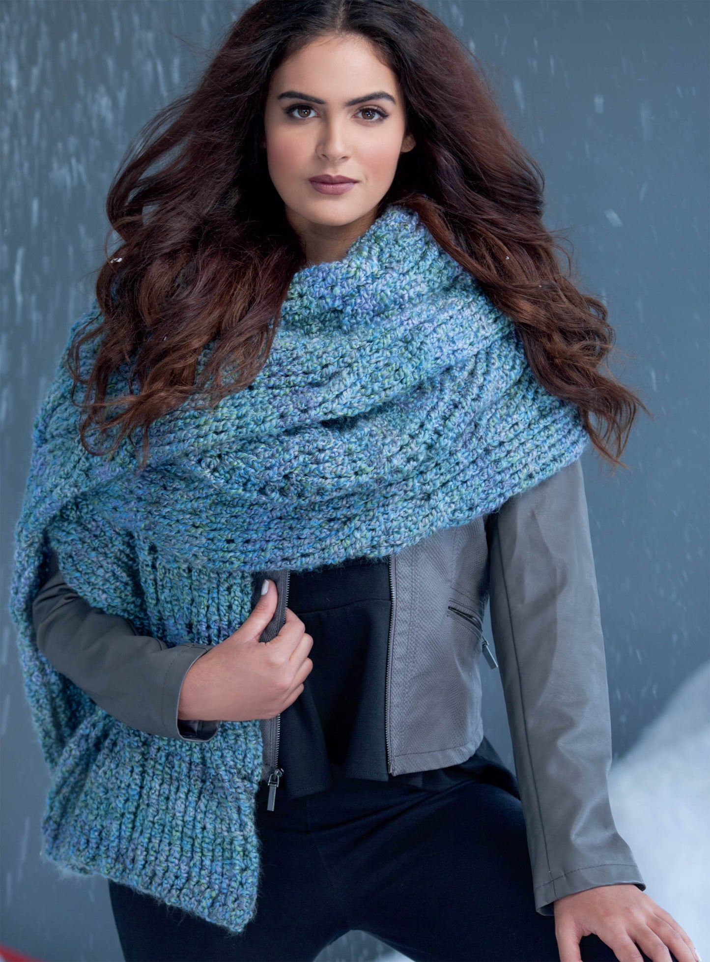 A woman with long wavy brown hair models a chunky blue scarf made using Leisure Arts' Super Scarves to Crochet Digital Download, draped over her shoulders with a gray jacket and black pants, standing in a snowy winter scene.
