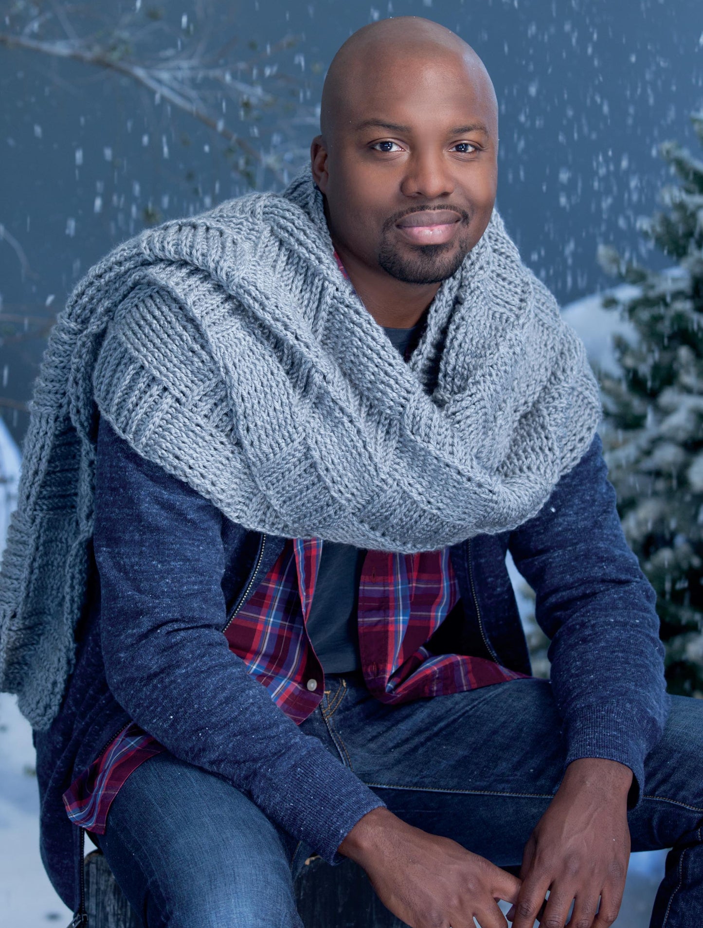 A man sits outdoors in the snow, wearing a chunky gray scarf made with patterns from the Super Scarves to Crochet Digital Download by Leisure Arts. He pairs it with a denim jacket, plaid shirt, and jeans among pine trees and falling snowflakes.