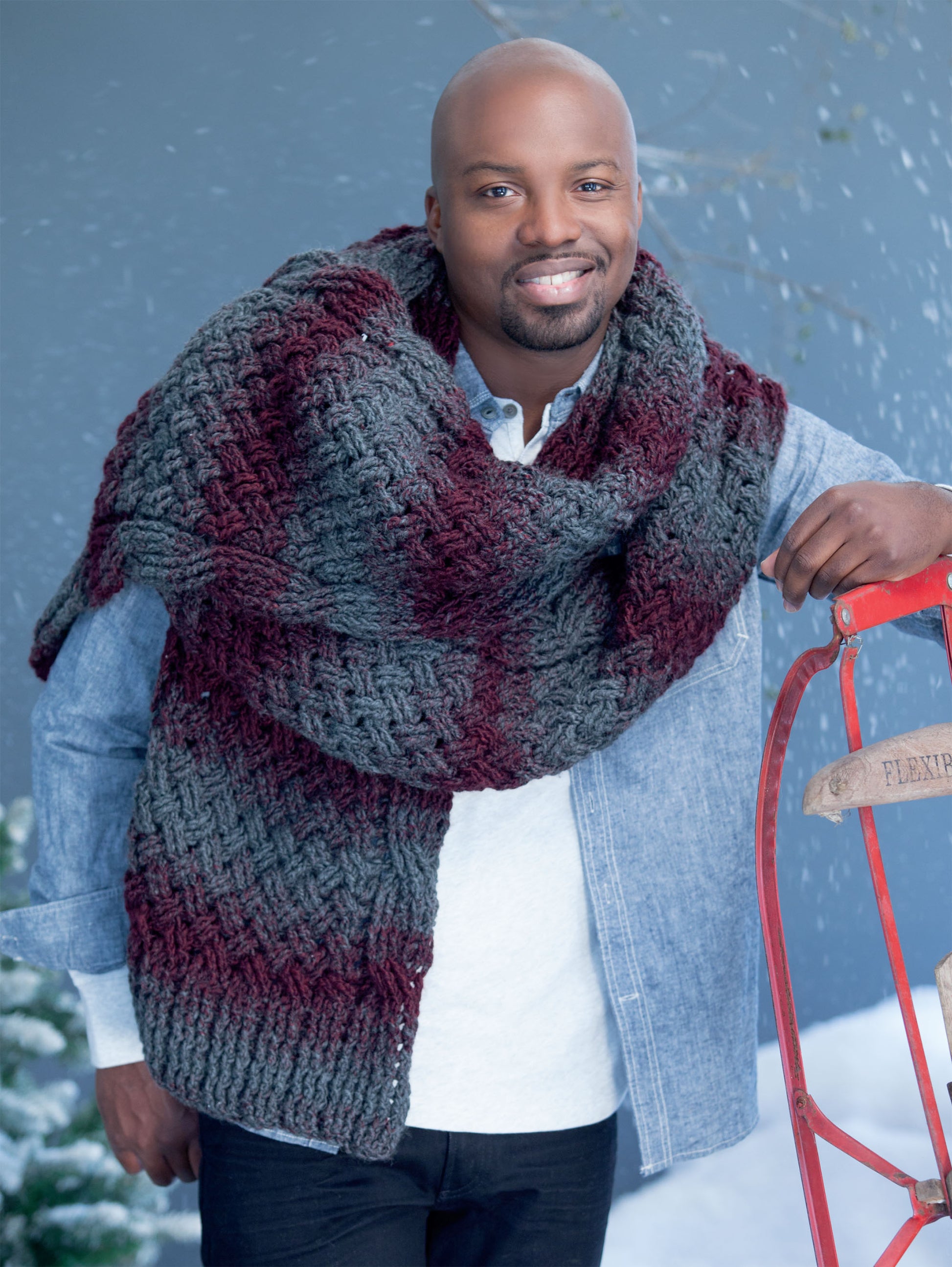 A smiling man in a blue denim shirt and maroon-and-gray scarf, from Leisure Arts’ Super Scarves to Crochet Digital Download, stands by a red sled in the snow—ideal inspiration for beginner crochet.