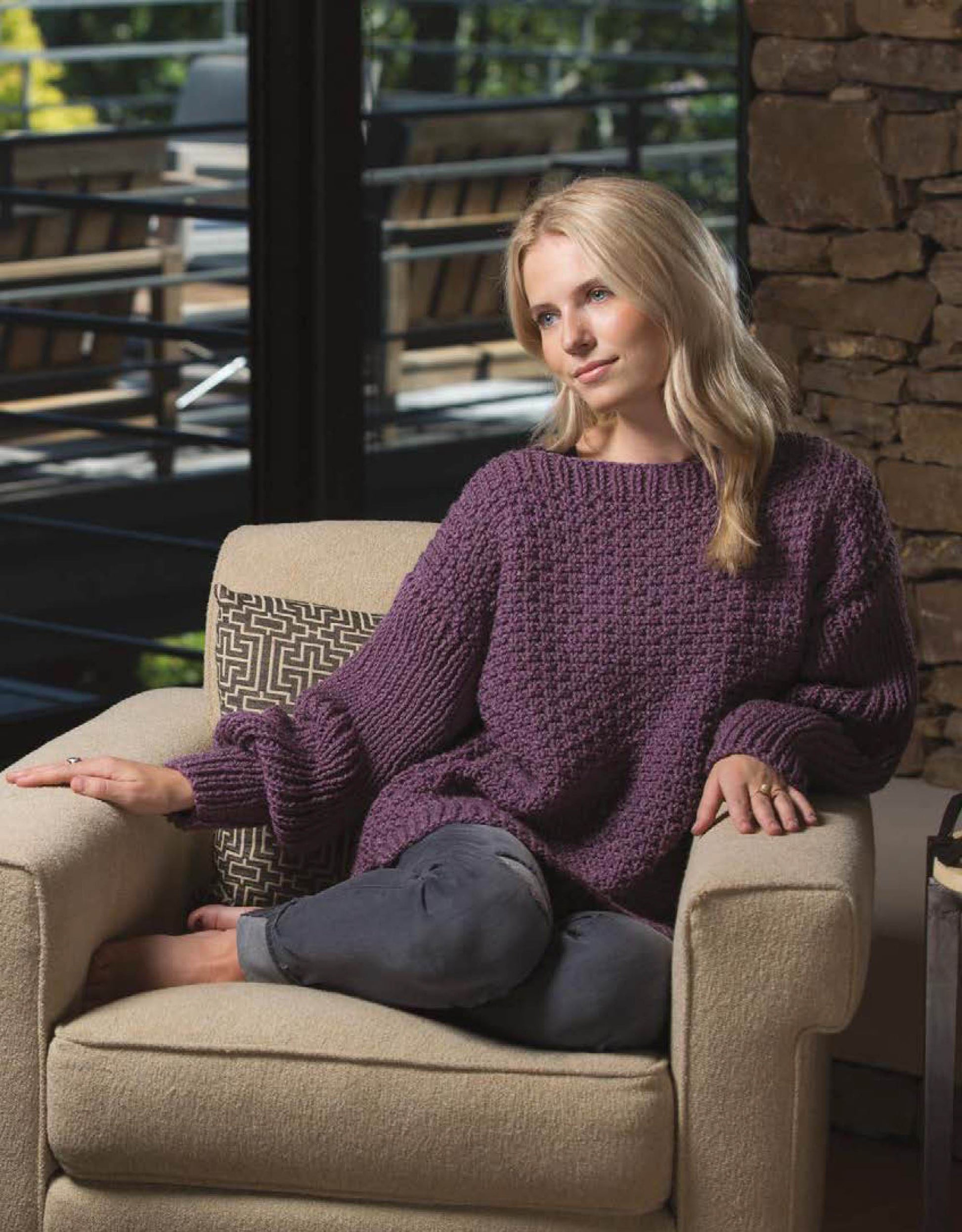 A woman with long blonde hair sits barefoot on a beige armchair, wearing a loose purple knit sweater—ideal inspiration for Leisure Arts Knit Cardigans projects—with her legs crossed and a patterned pillow beside her.