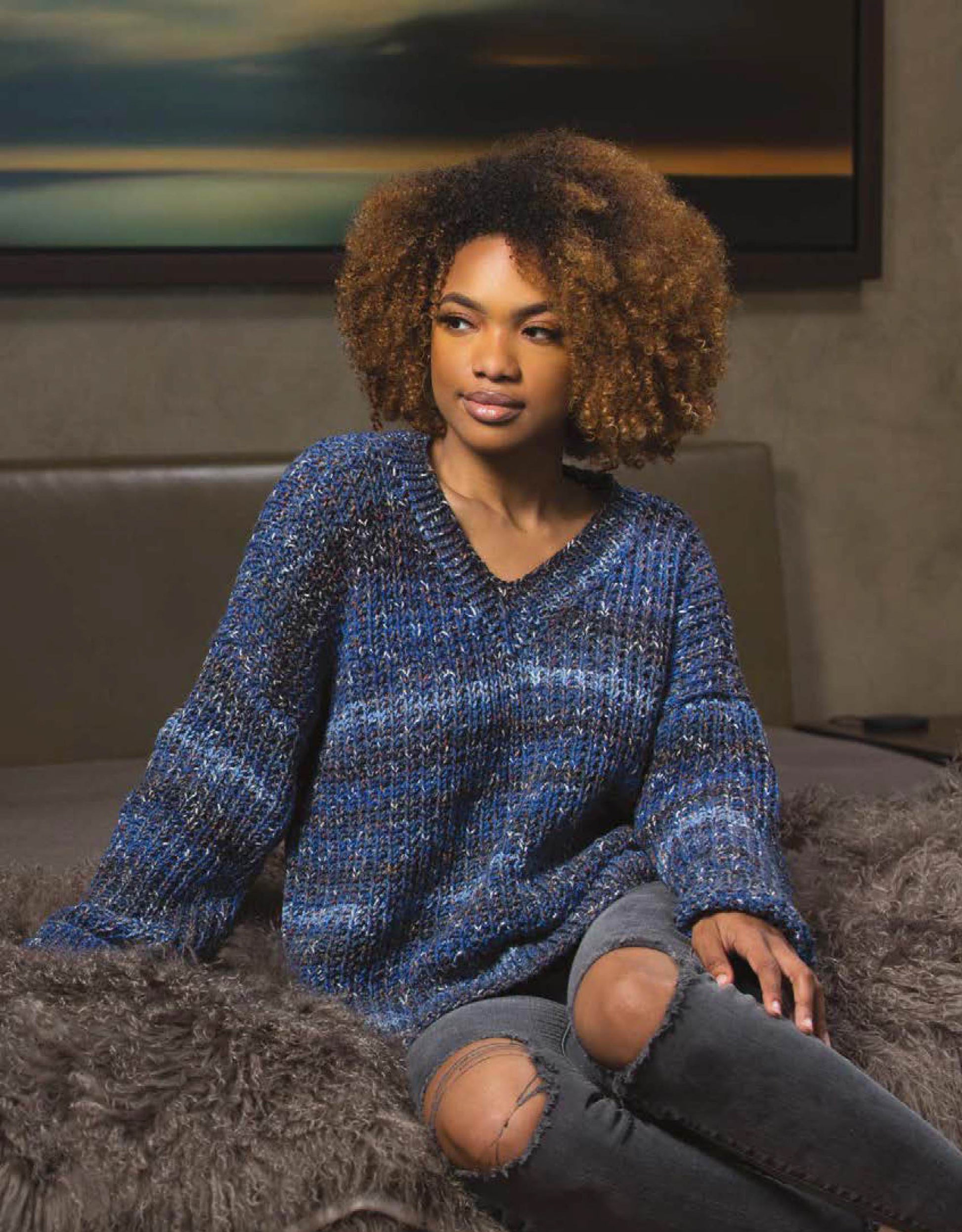 A woman with curly hair wears a chunky blue knit sweater inspired by Leisure Arts Knit Cardigans, sitting on a fur-covered surface and gazing thoughtfully in a softly lit room with art in the background.