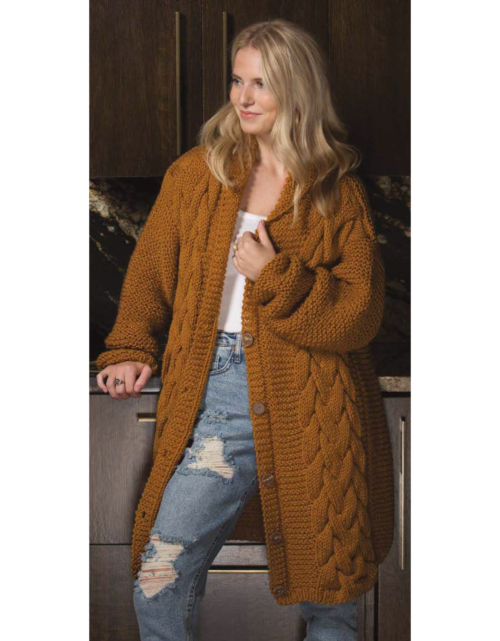 A woman smiles and looks to the side, wearing a chunky brown Knit Cardigan from Leisure Arts over a white top and ripped blue jeans, standing in front of dark wooden cabinets—great inspiration for cardigans and intermediate knitting projects.