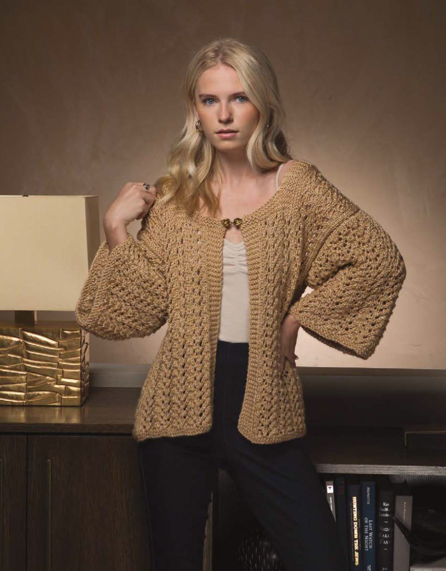 A woman with long blonde hair wears a textured tan Knit Cardigan by Leisure Arts over a light top and black pants, standing indoors beside a modern lamp and bookshelf—a cozy inspiration for sweater knitters.