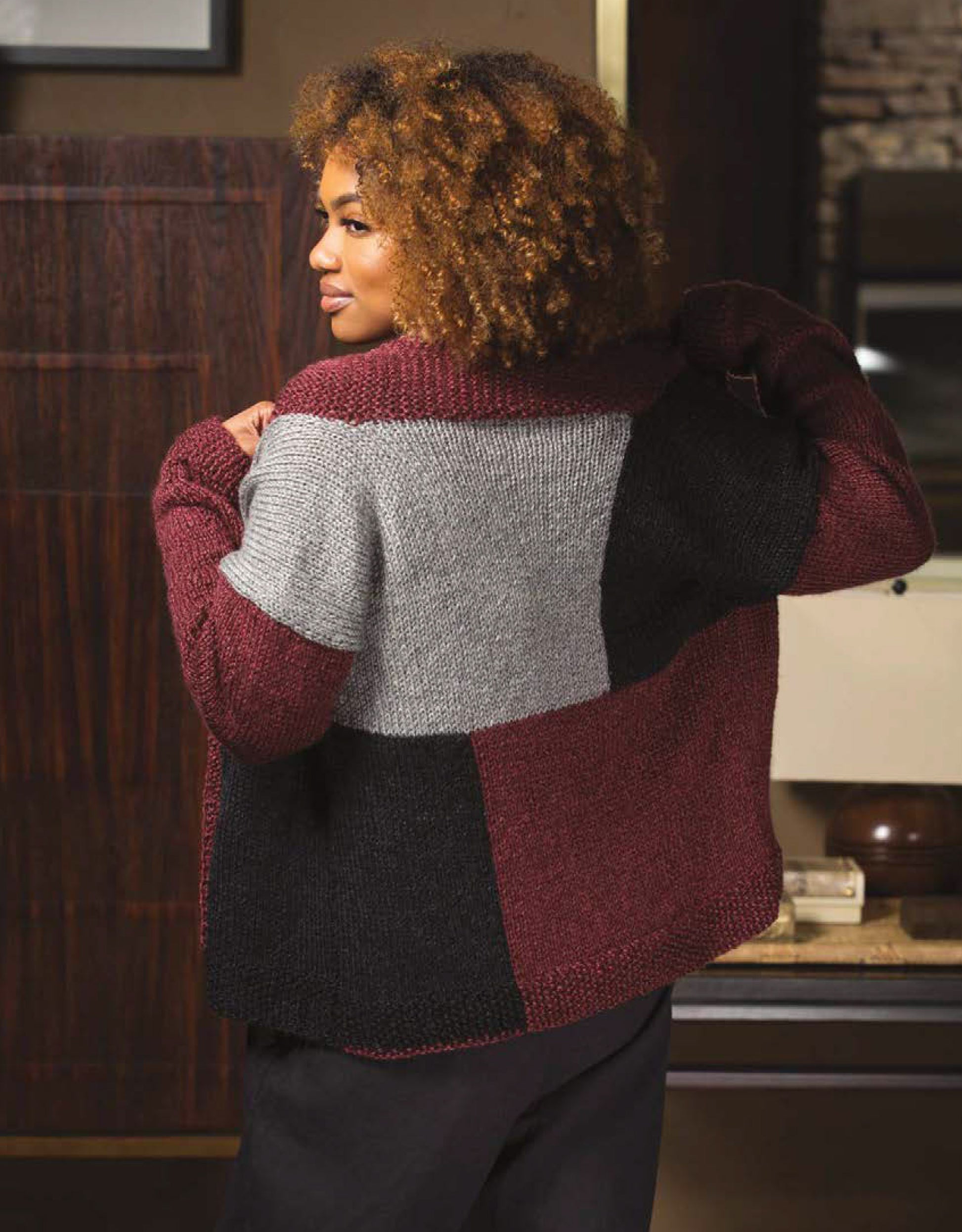 A person with curly hair wears the Leisure Arts Knit Cardigans in a burgundy, gray, and black color-blocked design, perfect for those shopping for women's cardigans or searching for sweater knitting pattern inspiration.