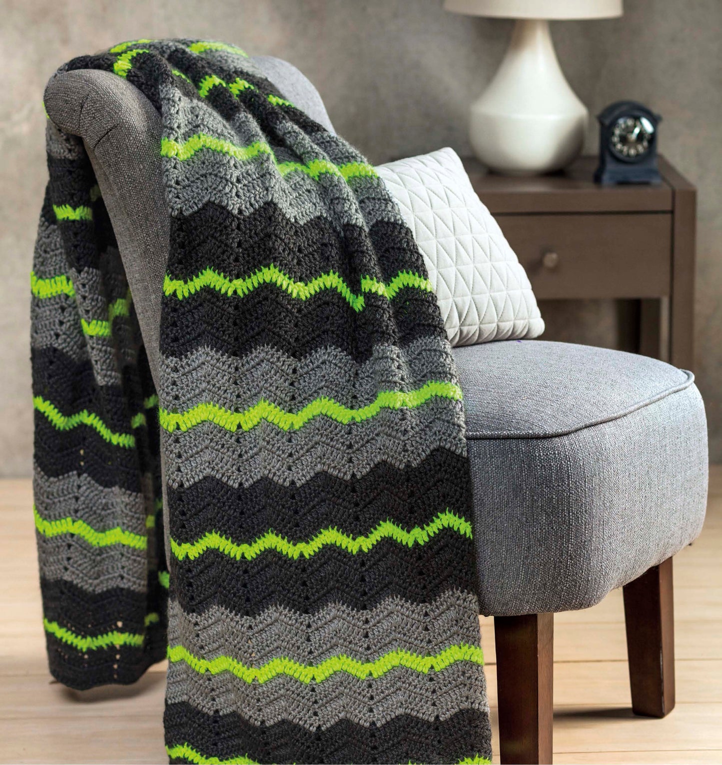 The "Shades of Grey Crochet" blanket by Leisure Arts, with black, gray, and bright green zigzag stripes, is draped over a gray chair with a white cushion. A wooden nightstand with a lamp and clock sits in the background.