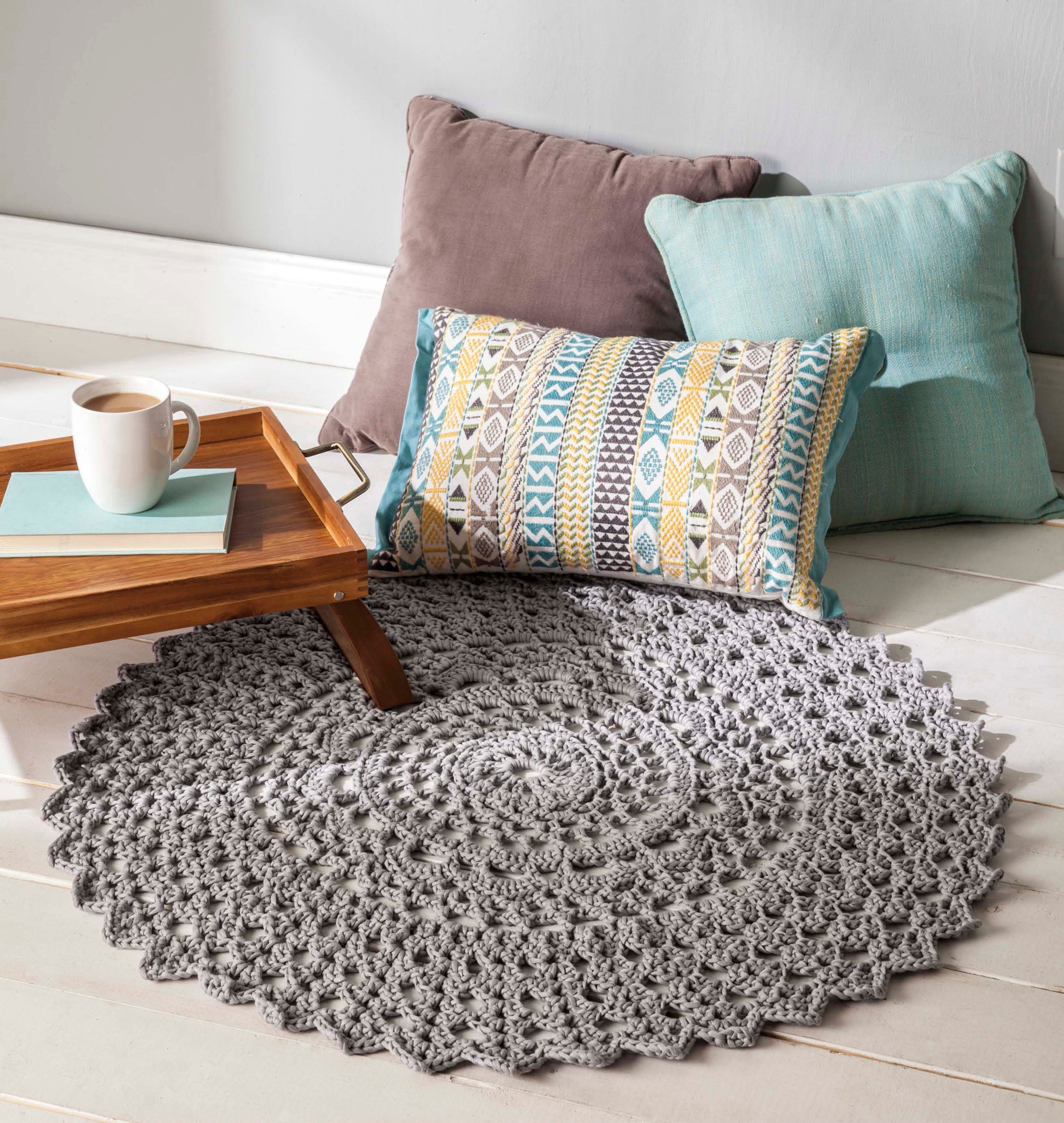 A cozy scene featuring three pillows, the "Shades of Grey Crochet" round rug by Leisure Arts showing off beautiful crochet textures, and a wooden tray with a mug and book.