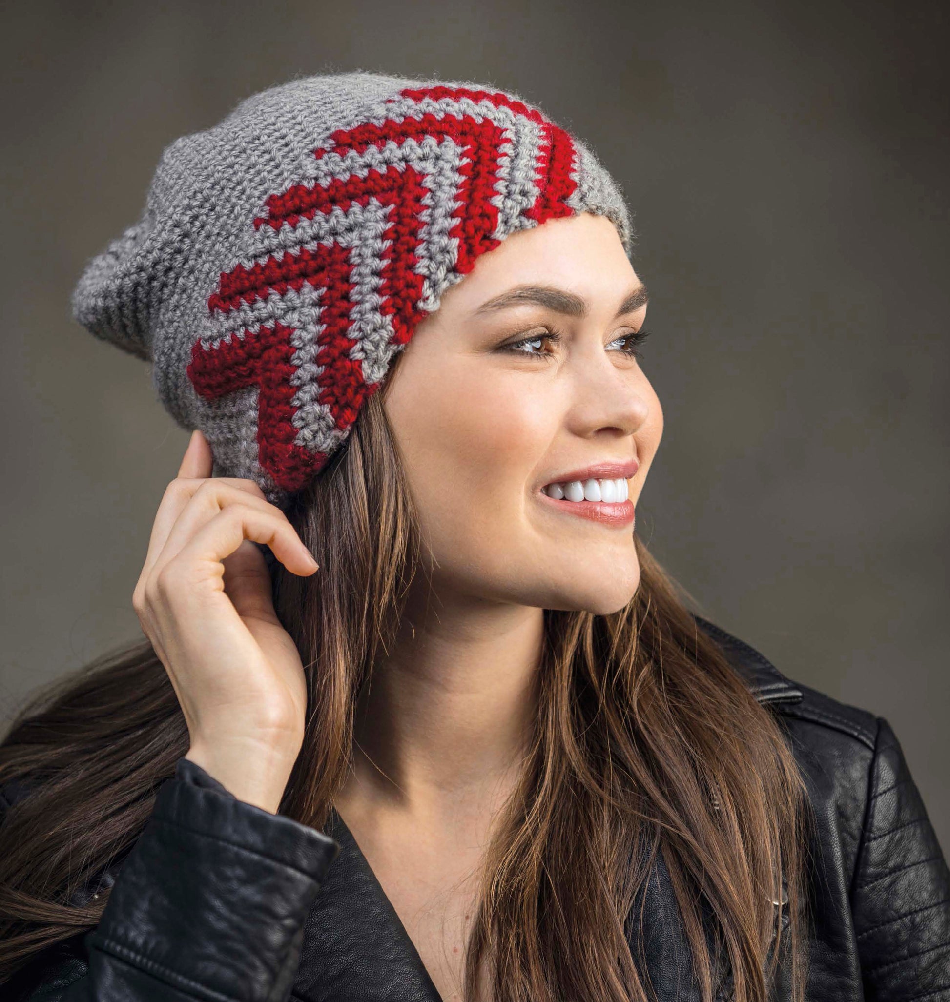 A smiling woman models the Leisure Arts "Shades of Grey Crochet" beanie, featuring red and dark gray zigzag patterns, rich crochet textures, and a stylish look paired with a black leather jacket.