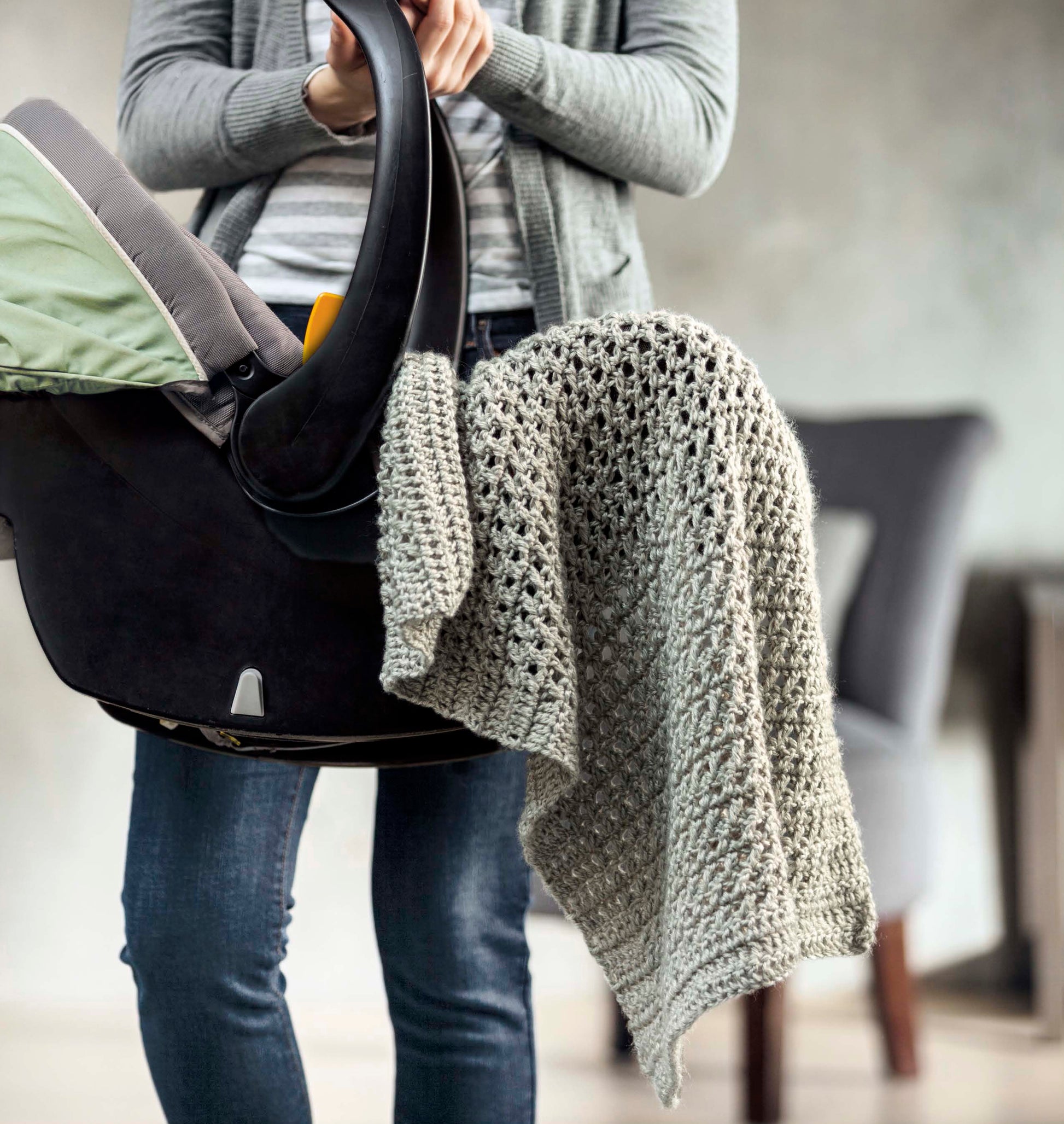 A person in jeans and a gray sweater holds a baby car seat draped with the Leisure Arts "Shades of Grey Crochet" blanket, highlighting its beautiful textures; the softly blurred background features a visible chair.