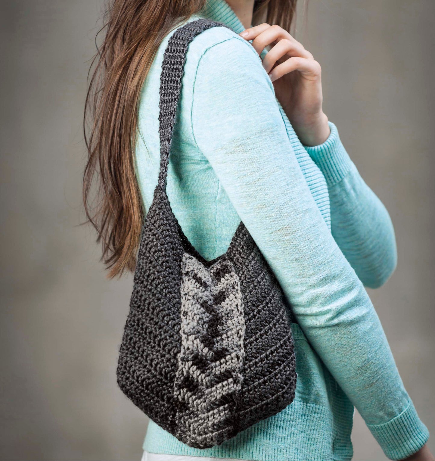 A woman in a light blue sweater holds the Leisure Arts "Shades of Grey Crochet" handmade shoulder bag, featuring dark and light gray chevron patterns and beautiful crochet textures against a neutral, blurred background.