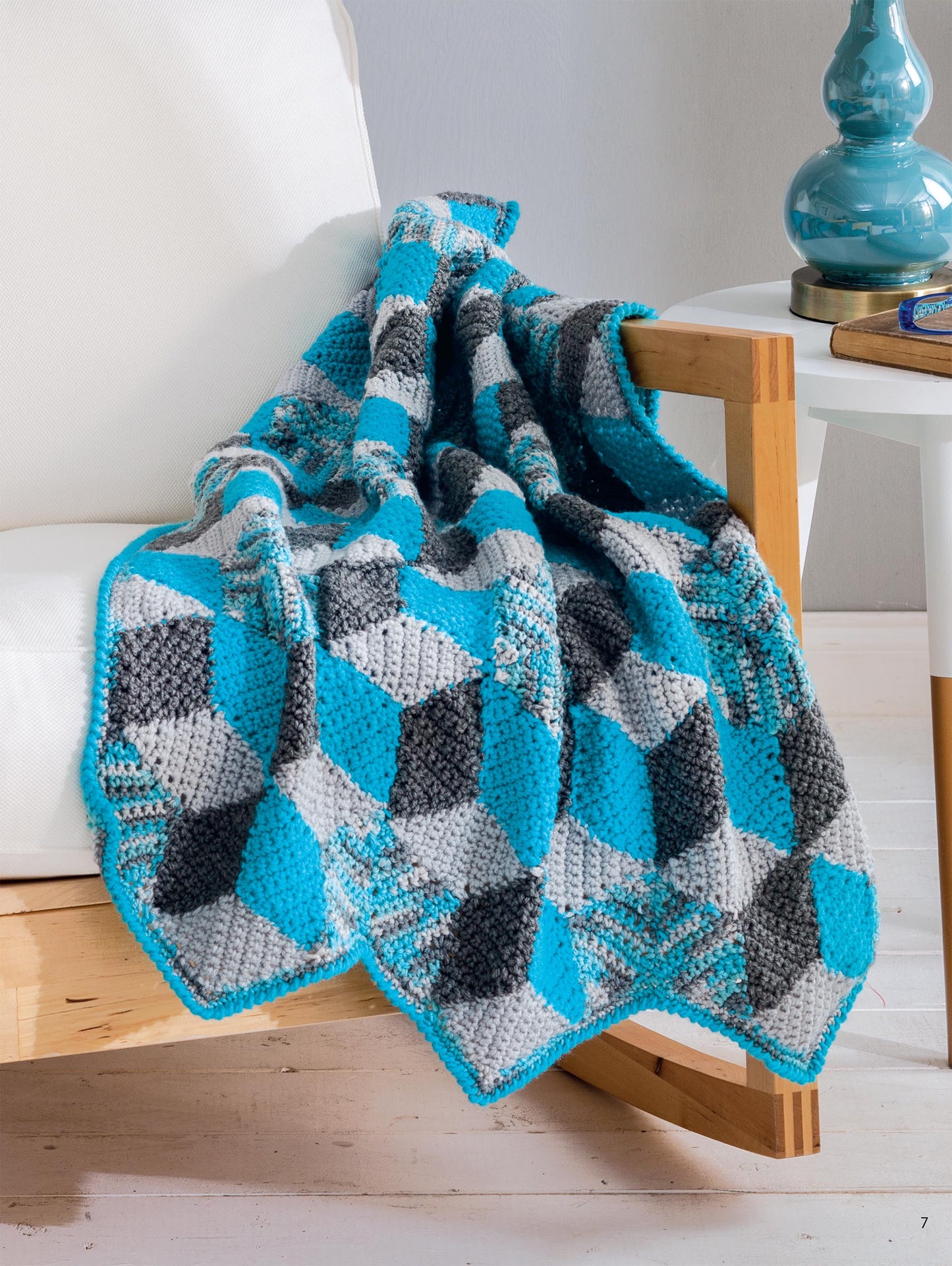 The Hooked On Quilted Crochet Digital Download by Leisure Arts features a geometric blue, gray, and black blanket draped over a wooden armchair, adding stylish decor beside a white side table topped with a blue lamp.