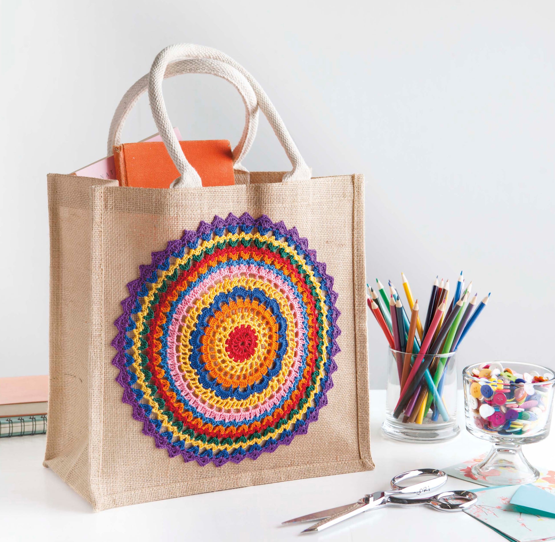 The Doilies Updated tote bag by Leisure Arts, featuring a colorful crocheted mandala design, sits on a white desk with craft supplies. The beige bag holds a closed orange book, perfect for storing your versatile yarn projects.
