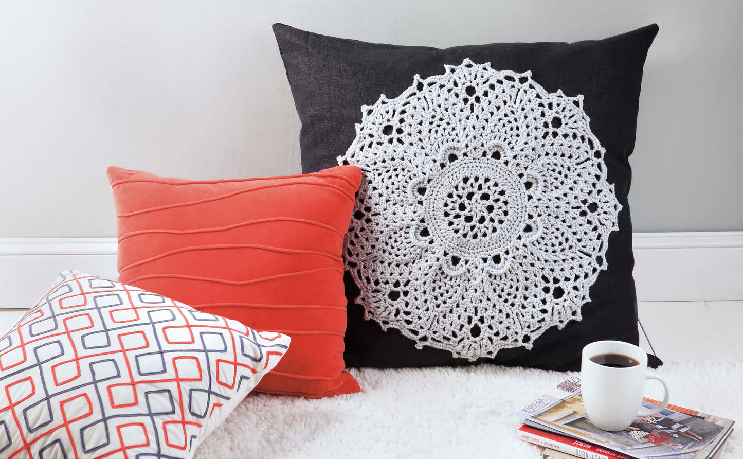 Three pillows rest on a white rug: one is the large black 'Doilies Updated' by Leisure Arts with a crochet design, another is coral with pleats, and the third has geometric patterns. Nearby are a cup of coffee and magazines.