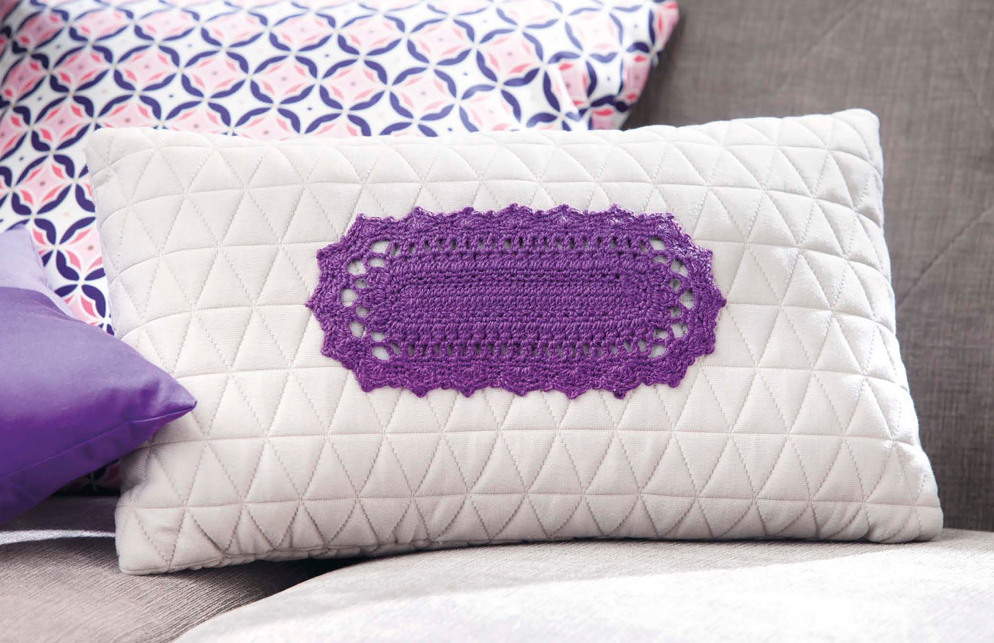 The Doilies Updated pillow by Leisure Arts—a white quilted pillow with a purple crocheted oval design—adds charm to your couch, beautifully complemented by patterned and solid purple pillows in the background.