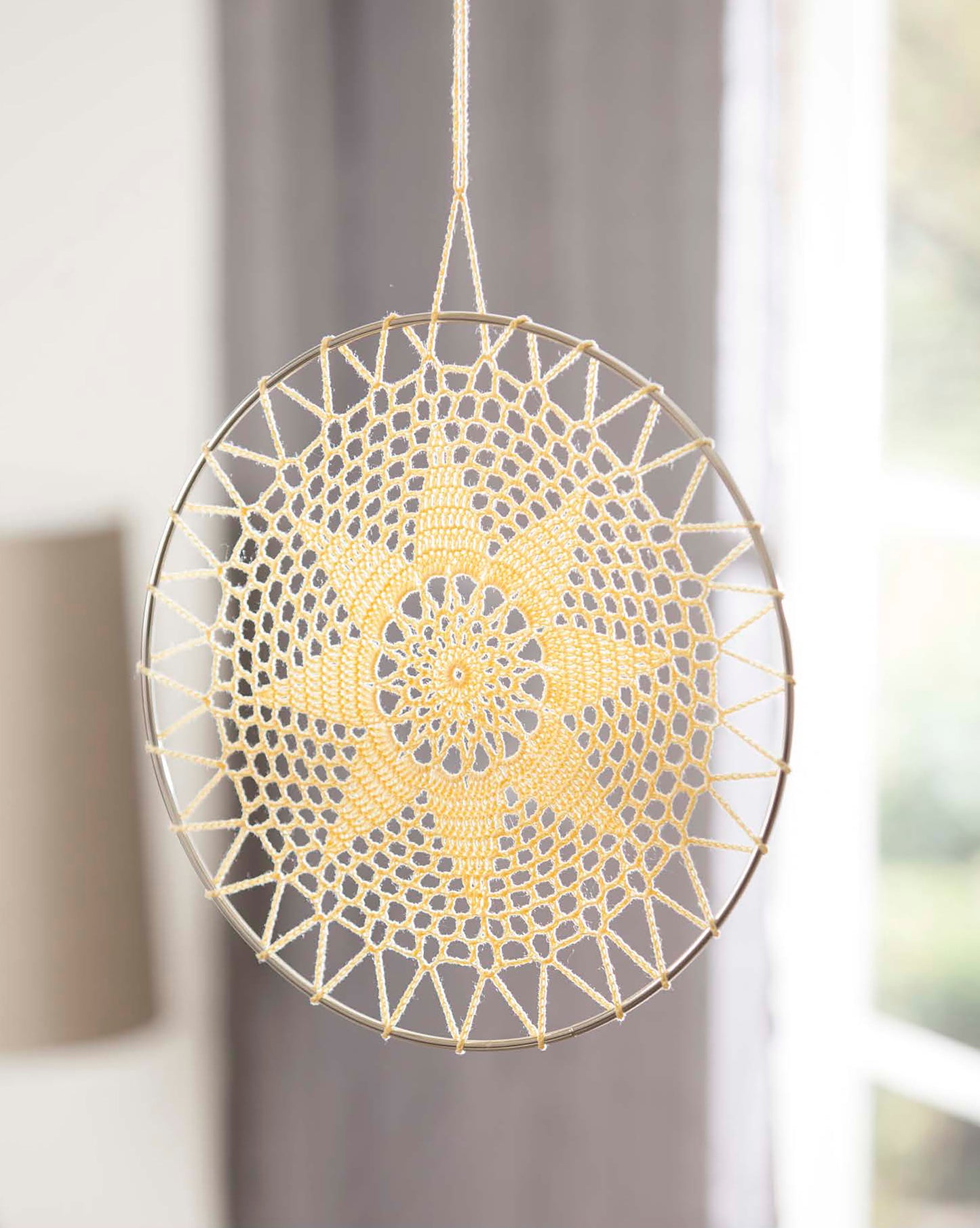 The Doilies Updated by Leisure Arts is a handmade dreamcatcher featuring a yellow crochet star and circular lace patterns, beautifully displayed with soft natural light in front of a blurred gray curtain and window.