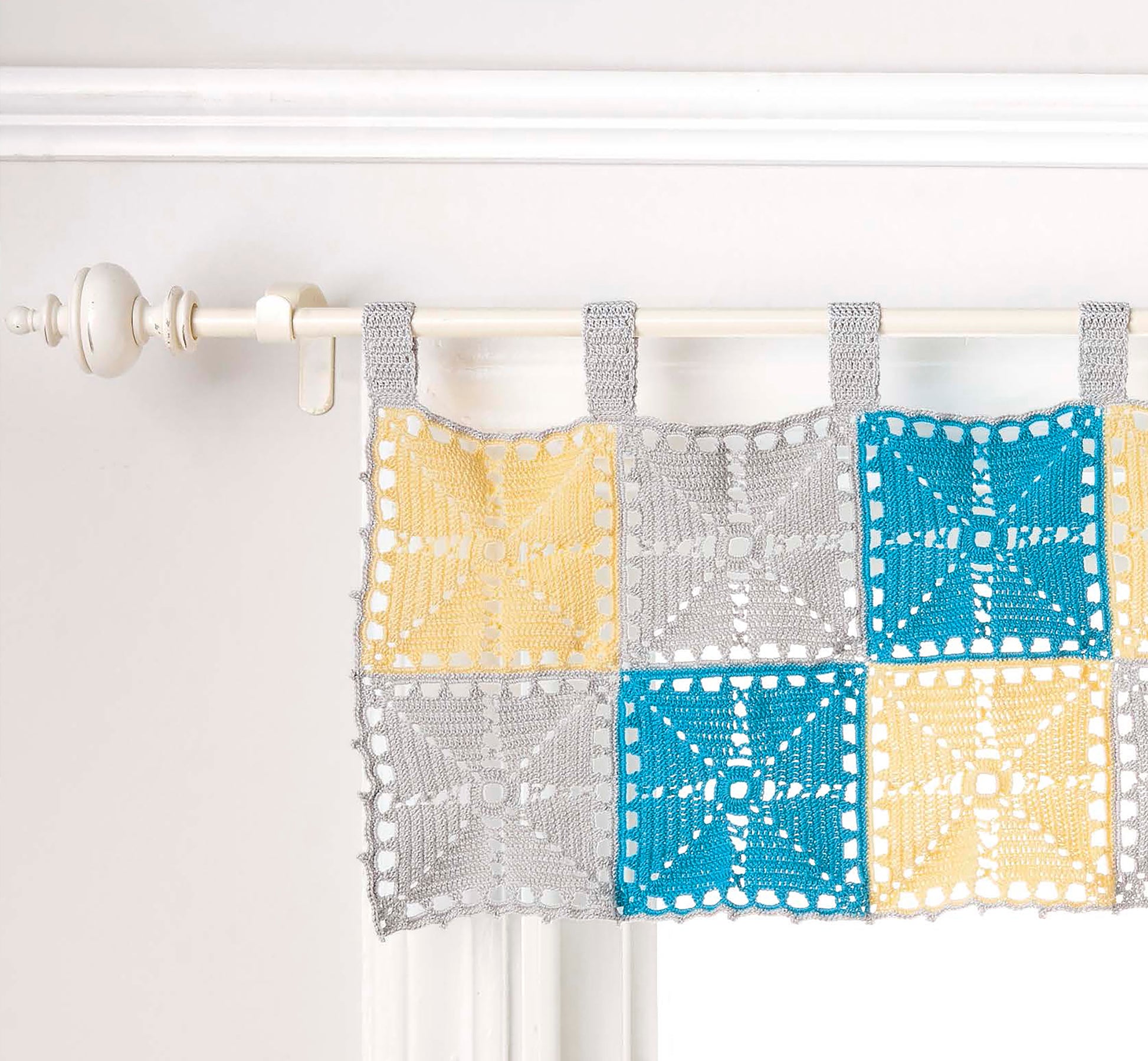 The Doilies Updated by Leisure Arts features a crochet curtain with square patterns in yellow, blue, and gray on a white rod—showcasing stylish home decor perfect for versatile yarn projects.