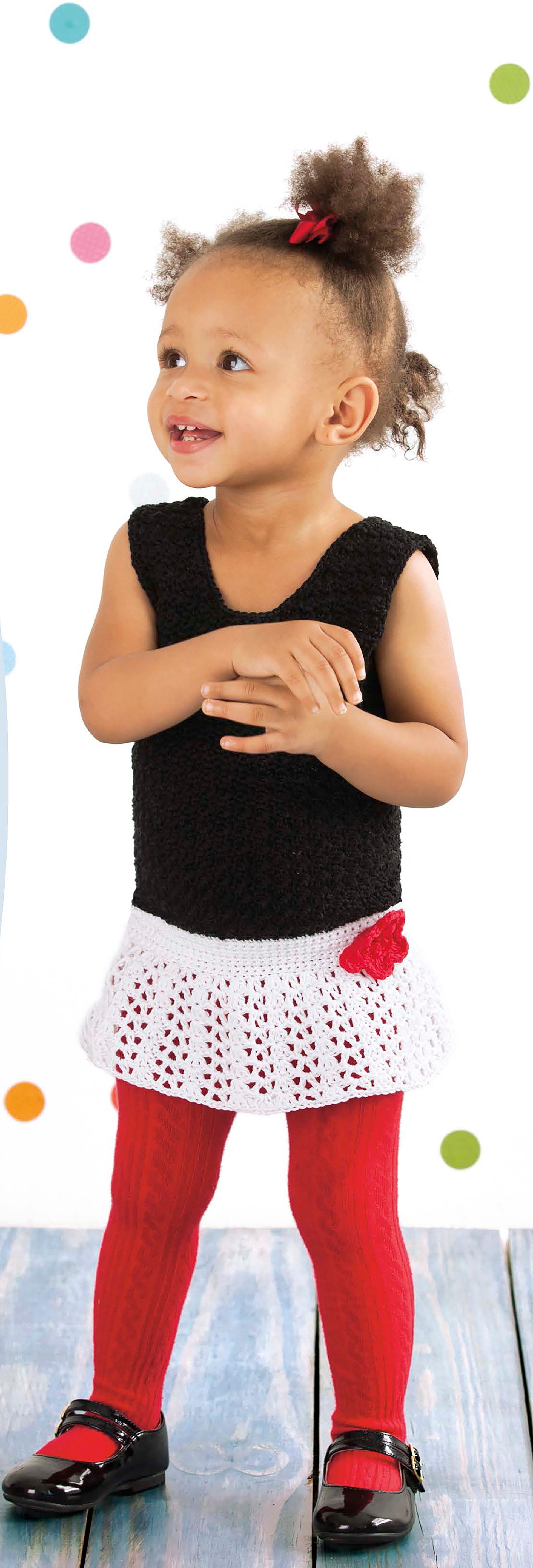 A young child models the Leisure Arts Lacy Little Girl Dress—a white crochet skirt with a red heart—paired with a black sleeveless top, red tights, and black shoes, standing on a wooden floor with colorful polka dot backdrop.