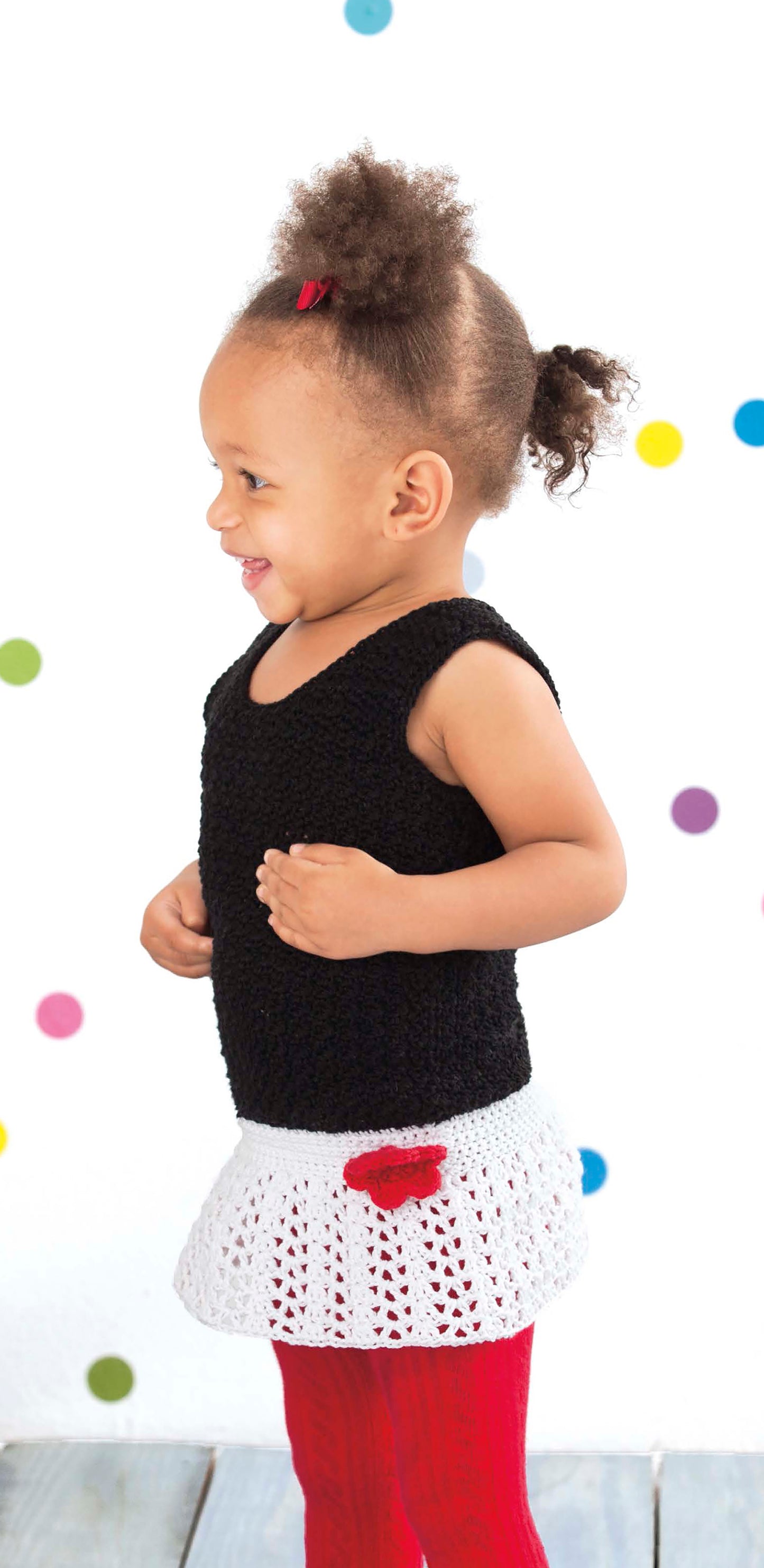 A happy toddler with curly hair in a Lacy Little Girl Dress by Leisure Arts—featuring a black sleeveless top, white crochet skirt, and red flower—stands before a white background with colorful polka dots.