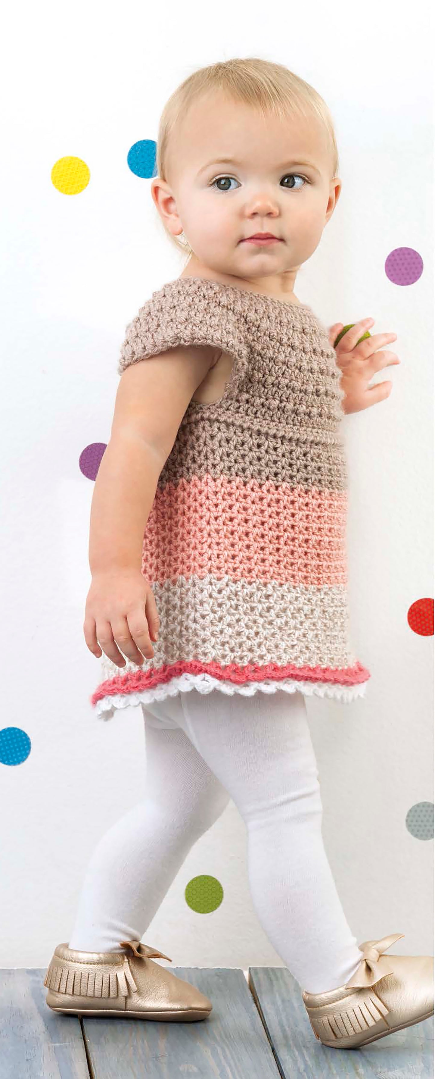 A young toddler with short blonde hair models the Lacy Little Girl Dresses by Leisure Arts, featuring brown, pink, and white crochet, paired with white leggings and gold shoes against a white wall with colorful polka dots.