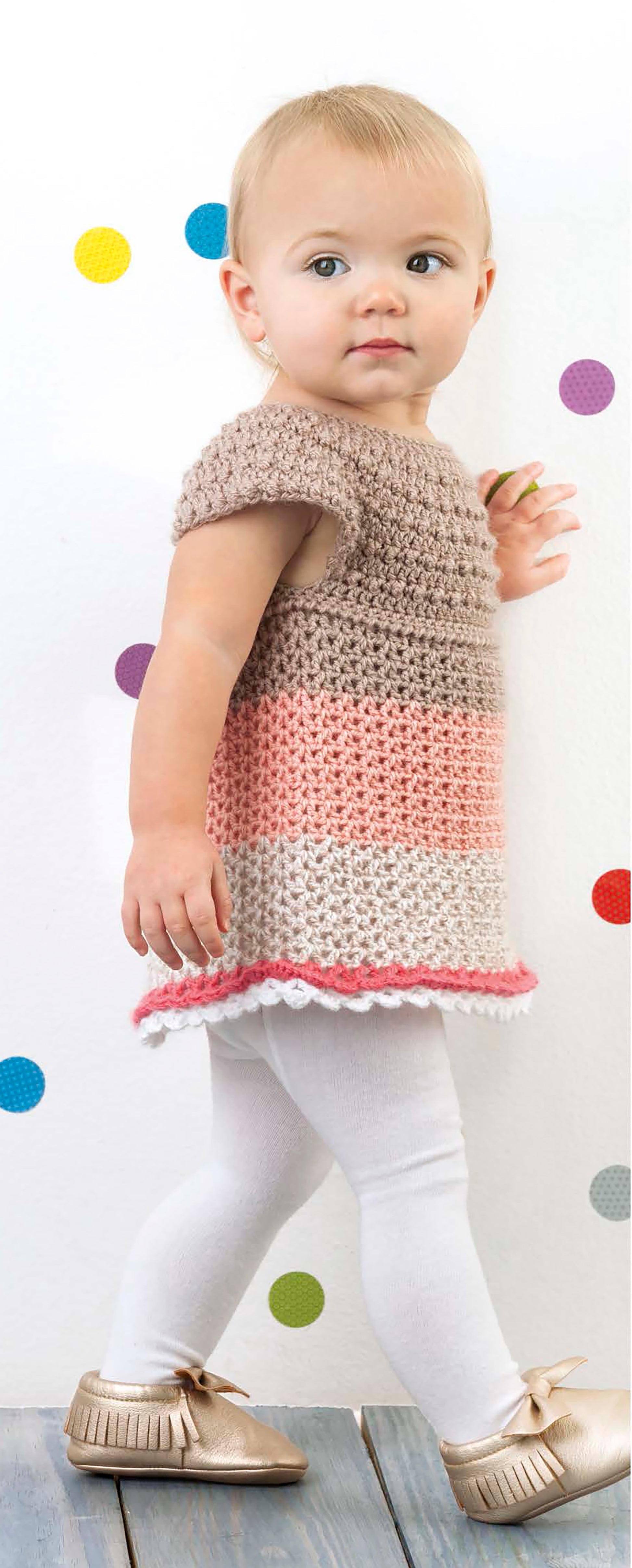 A young toddler with short blonde hair models the Lacy Little Girl Dresses by Leisure Arts, featuring brown, pink, and white crochet, paired with white leggings and gold shoes against a white wall with colorful polka dots.