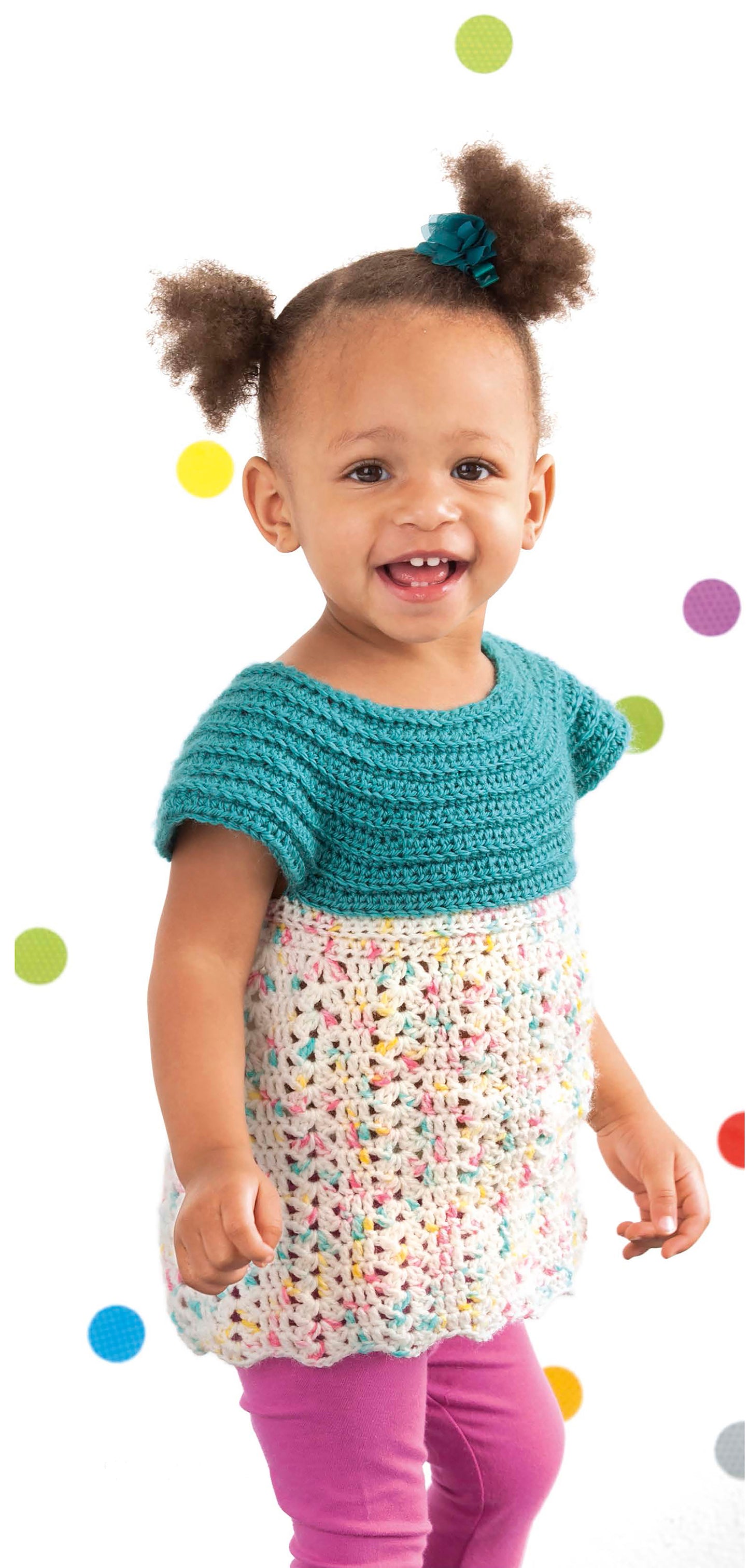 A smiling toddler with pigtails wears a Lacy Little Girl Dress by Leisure Arts, featuring a teal crochet top and multicolored skirt over pink leggings, standing before a white background with colorful polka dots.