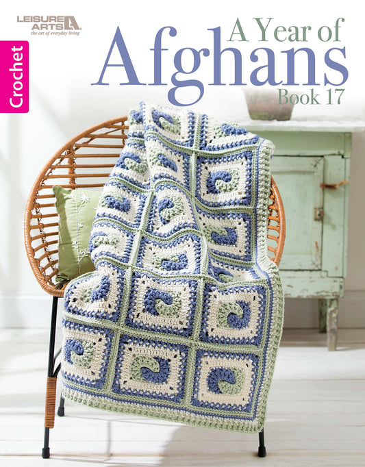 A crocheted afghan with blue, green, and white geometric patterns drapes over a wicker chair. "A Year of Afghans, Book 17 Digital Download" by Leisure Arts is featured, with a green side table and potted plant in the background.