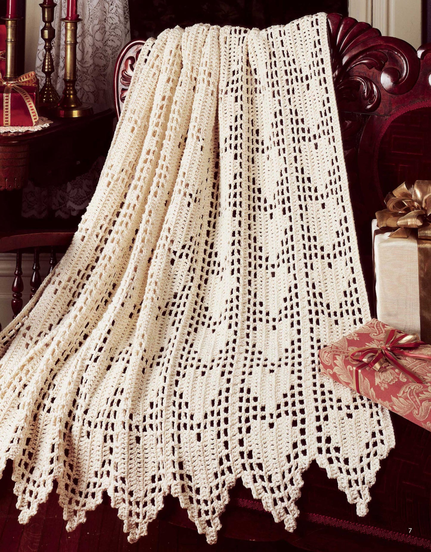 A cream-colored crocheted lace afghan from Leisure Arts’ "A Year of Afghans, Book 17 Digital Download" drapes over a carved wooden chair, with wrapped gifts and candles nearby for a cozy, festive atmosphere.