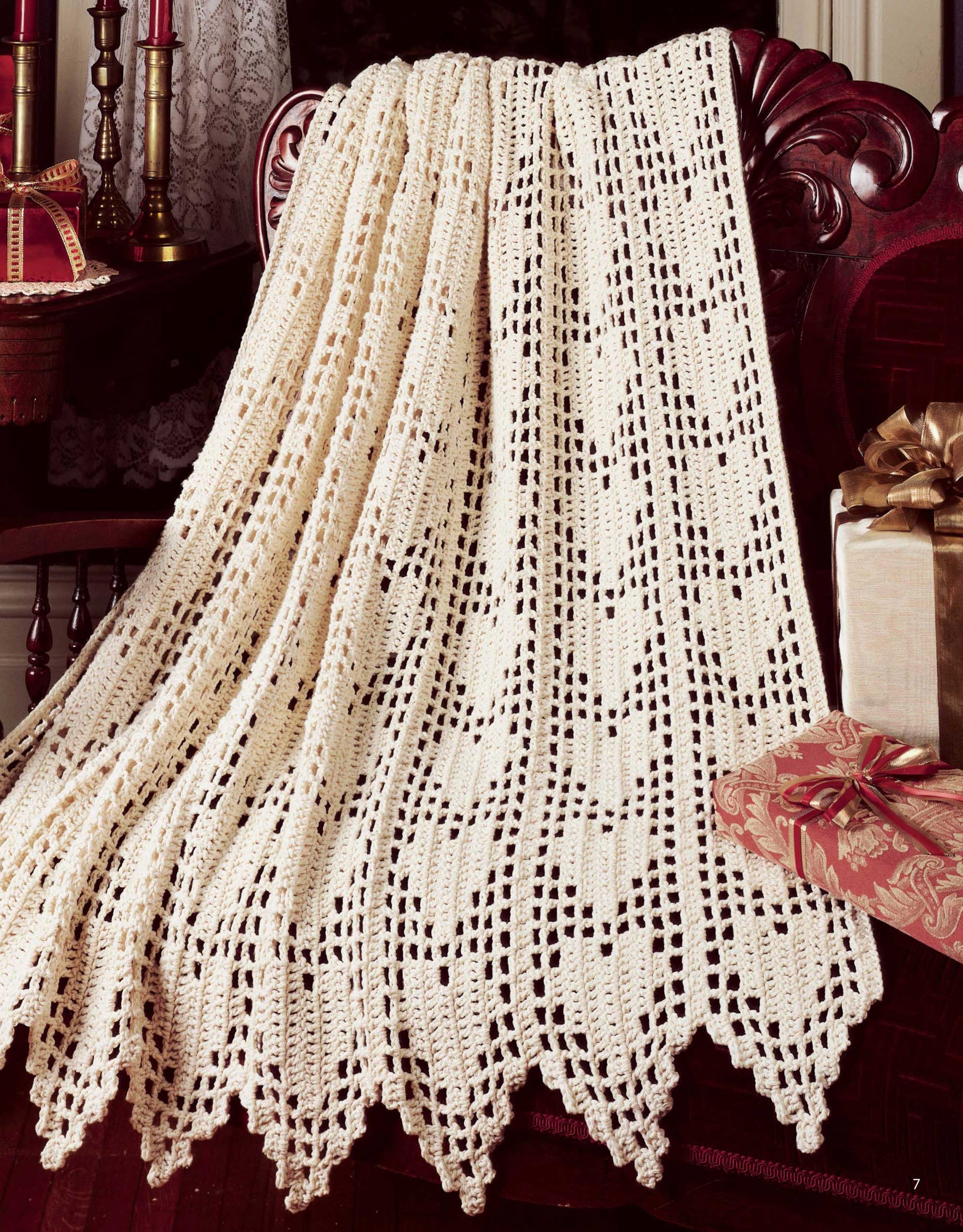 A cream-colored crocheted lace afghan from Leisure Arts’ "A Year of Afghans, Book 17 Digital Download" drapes over a carved wooden chair, with wrapped gifts and candles nearby for a cozy, festive atmosphere.