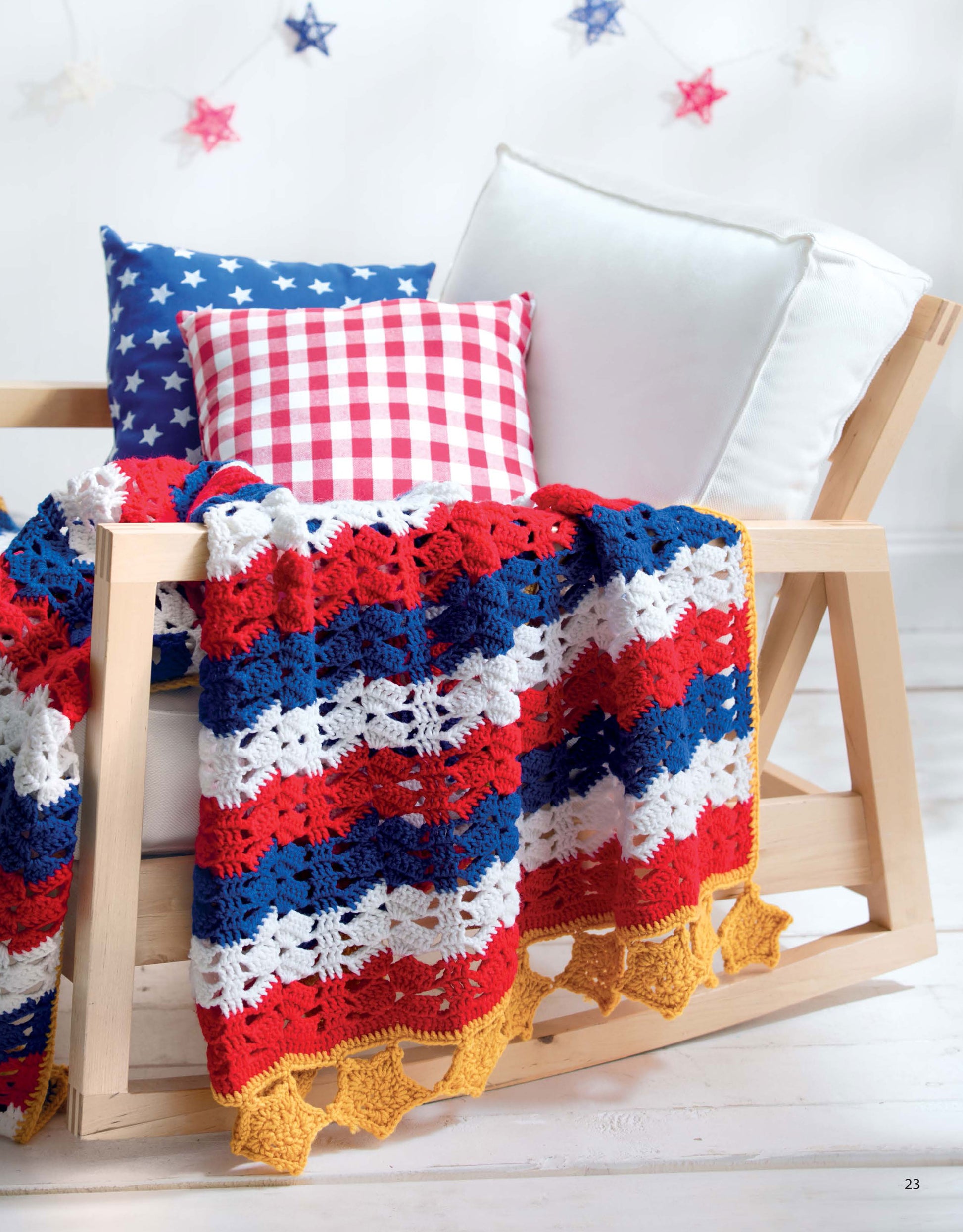 A wooden chair with star and checkered pillows is draped with a red, white, and blue striped afghan featuring a yellow star border from A Year of Afghans, Book 17 by Leisure Arts. Star garland hangs in the blurred background.