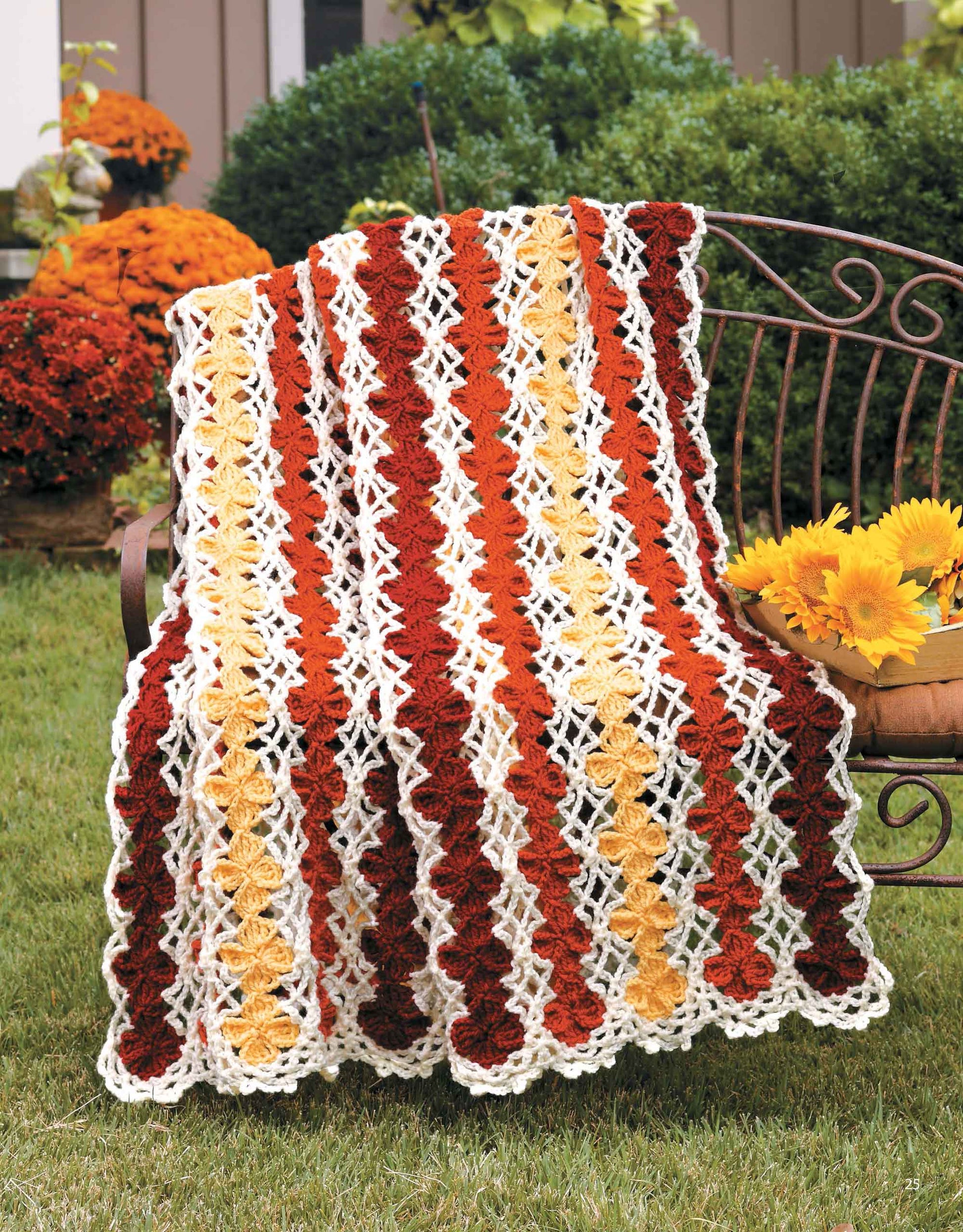 The "A Year of Afghans, Book 17" digital pattern by Leisure Arts features a red, orange, yellow, and white geometric crocheted blanket draped over an outdoor bench beside sunflowers and marigolds.
