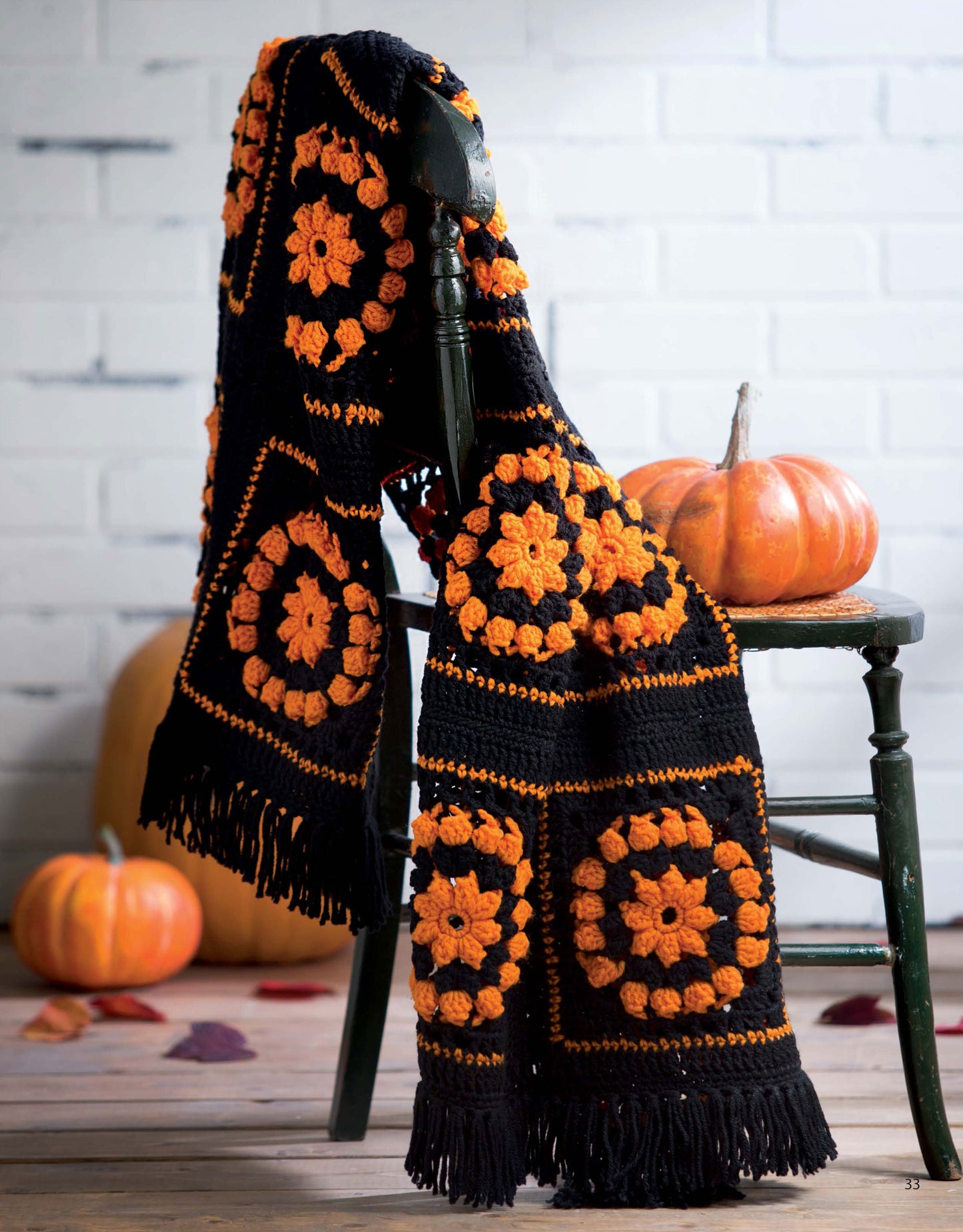 A crochet scarf from "A Year of Afghans, Book 17" by Leisure Arts hangs over a dark wooden chair beside pumpkins and autumn leaves on a wood floor, set against a white brick wall. Digital download available.