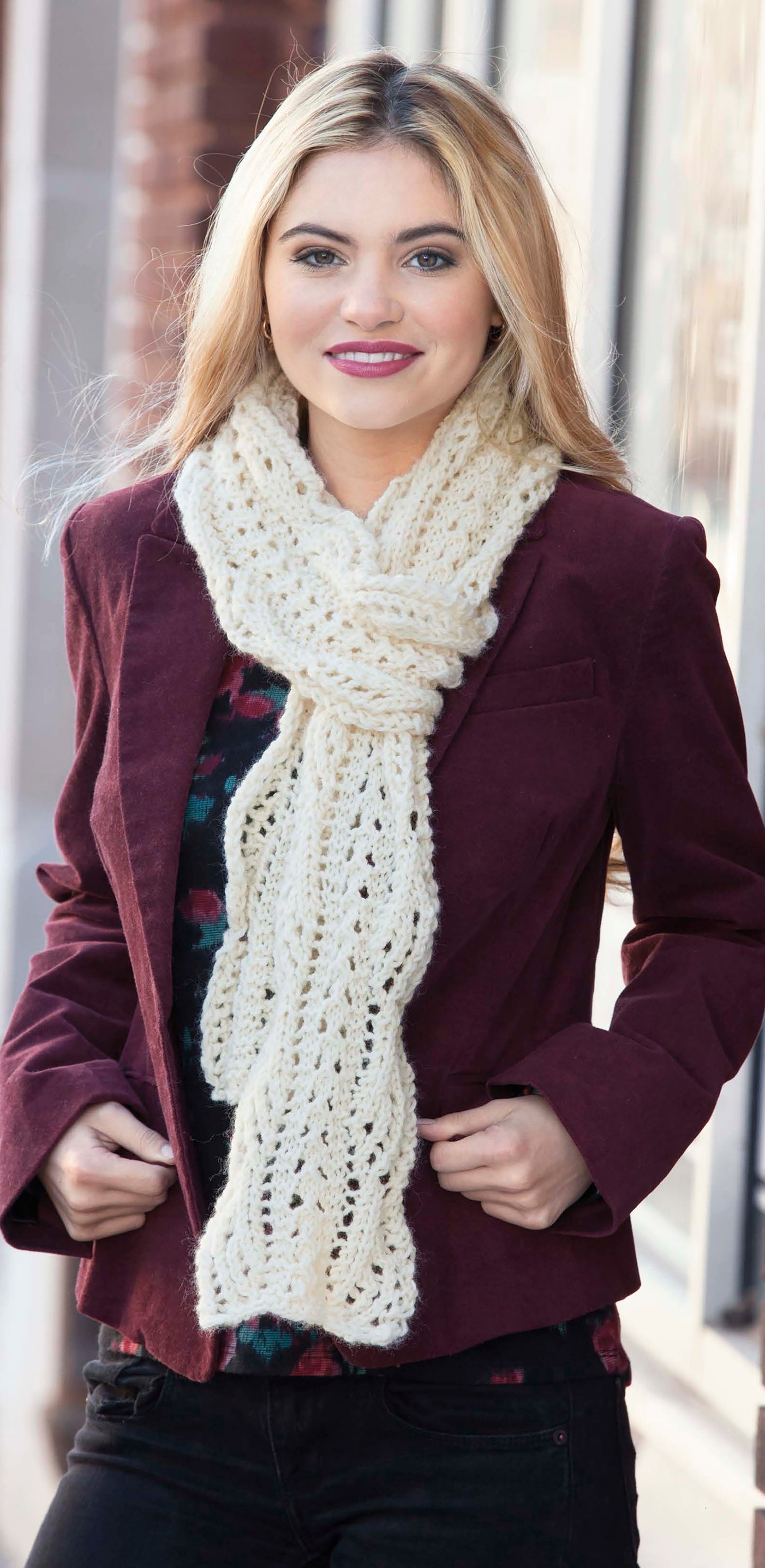 A woman with long blonde hair smiles at the camera outdoors, wearing a chunky cream-colored scarf made with super bulky yarn using the "Wild About Texture" pattern by Leisure Arts, paired with a burgundy jacket.