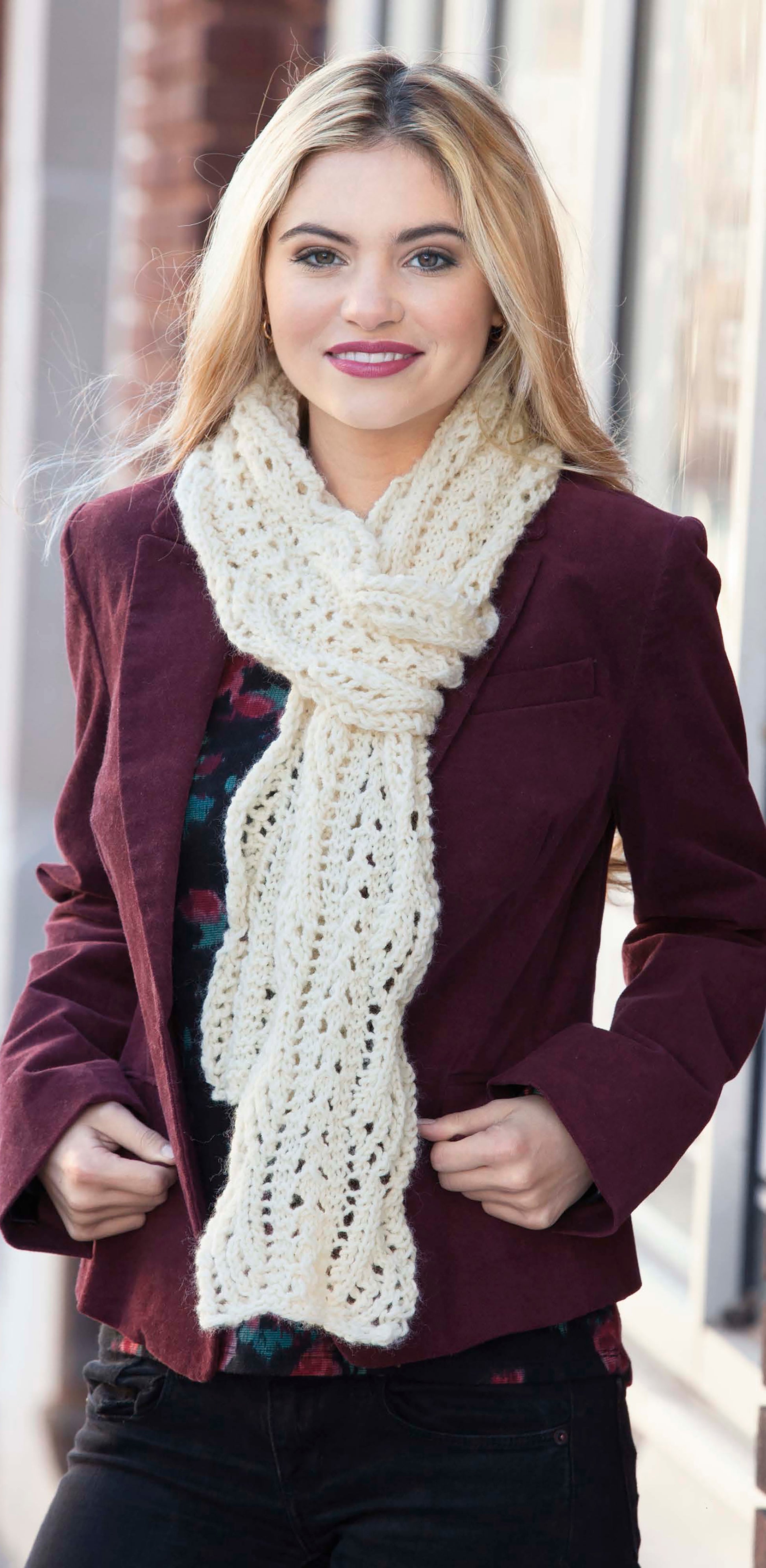 A woman with long blonde hair smiles at the camera outdoors, wearing a chunky cream-colored scarf made with super bulky yarn using the "Wild About Texture" pattern by Leisure Arts, paired with a burgundy jacket.