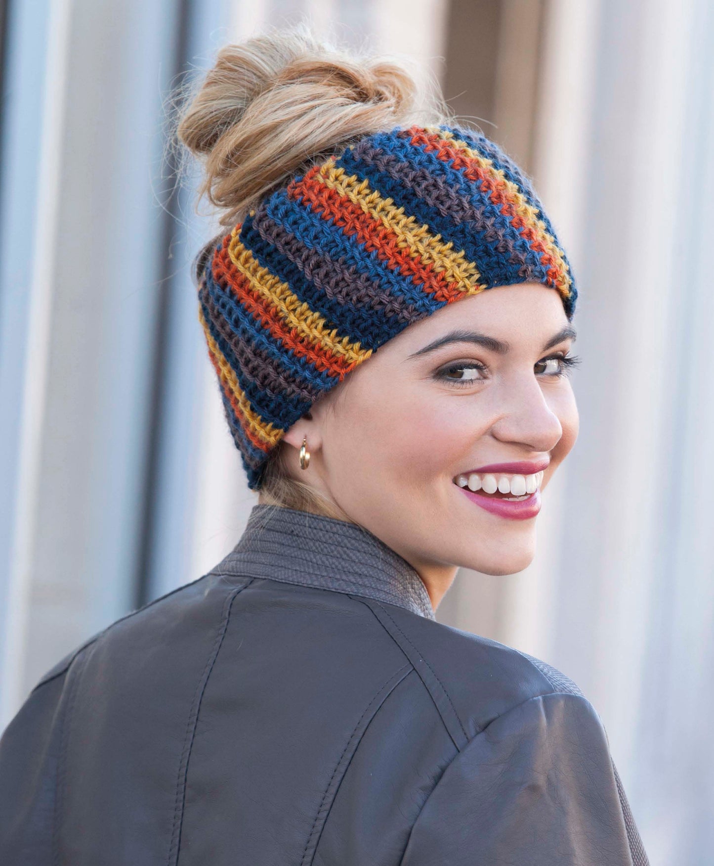 A smiling woman with light brown hair in a bun wears a colorful striped knit headband made with Wild About Texture by Leisure Arts, ideal for super bulky yarn projects. She pairs it with a gray jacket and looks over her shoulder outdoors.