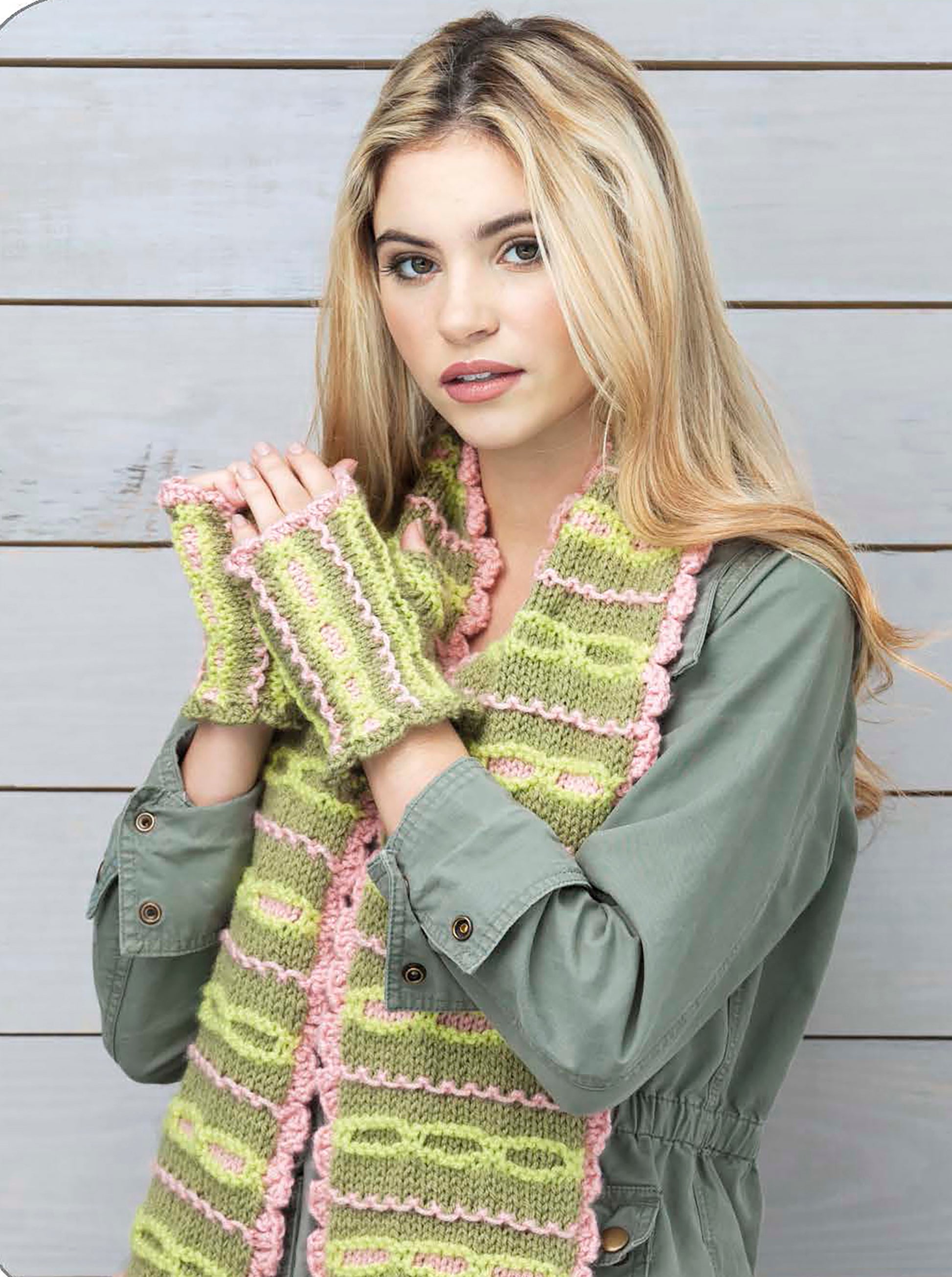 A young woman with long blonde hair wears the Wild About Texture scarf and matching fingerless gloves by Leisure Arts in green and pink, paired with a sage green jacket, standing before a light wooden background.