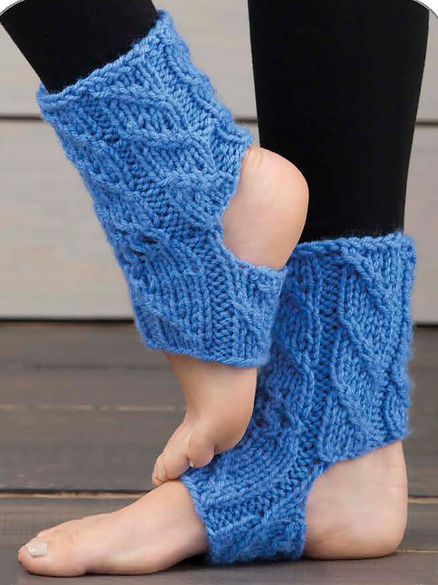 Close-up of feet in blue, knitted yoga socks—crafted from super bulky yarn—showing open heels and toes over black leggings on a wooden floor. Create your own with "Wild About Texture" by Leisure Arts, perfect for your next knit project!.