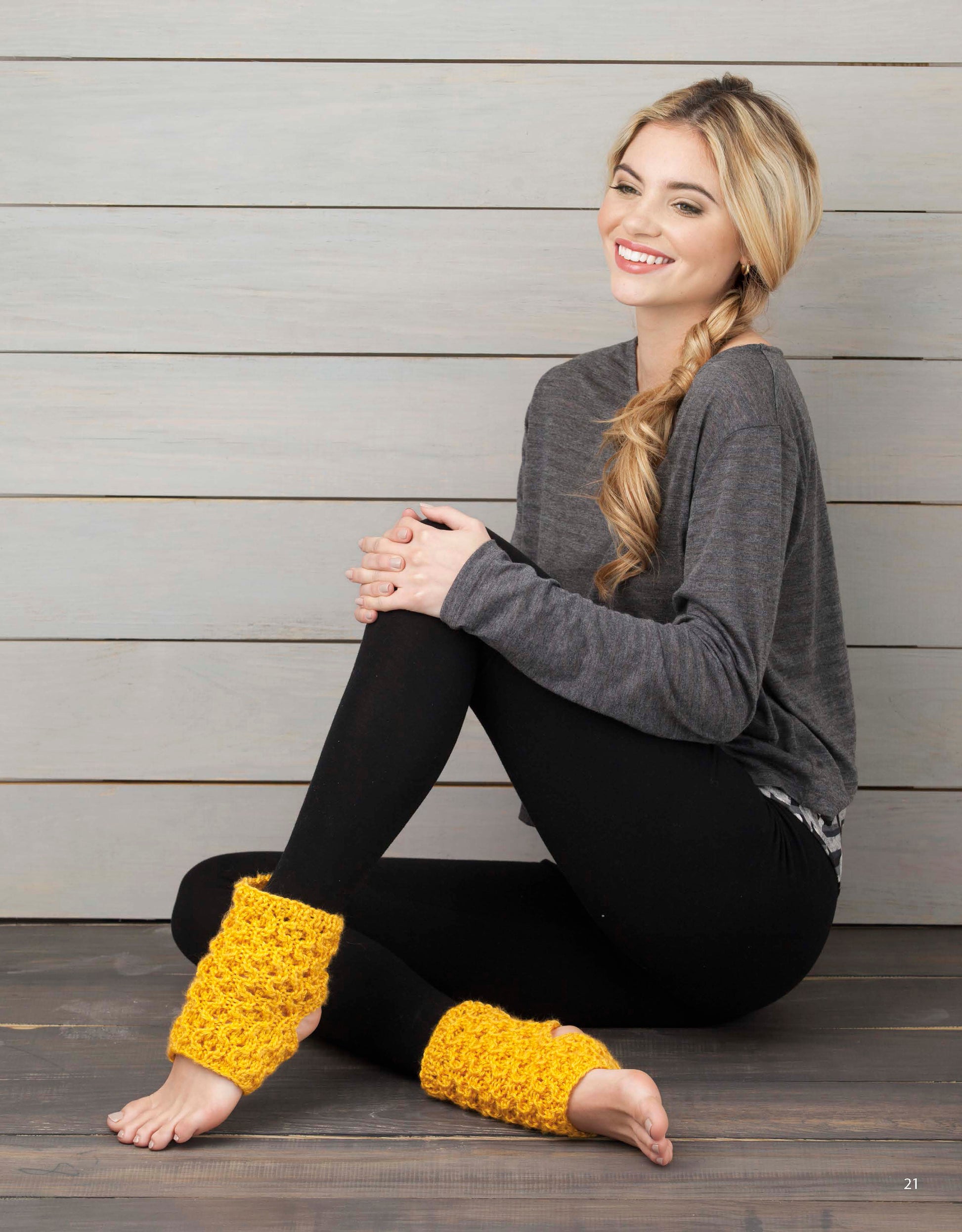 A smiling woman with long blonde hair in a side braid sits on the floor, wearing yellow leg warmers made with super bulky yarn from the Wild About Texture collection by Leisure Arts. She is dressed in a gray top and black leggings against a wood backdrop.