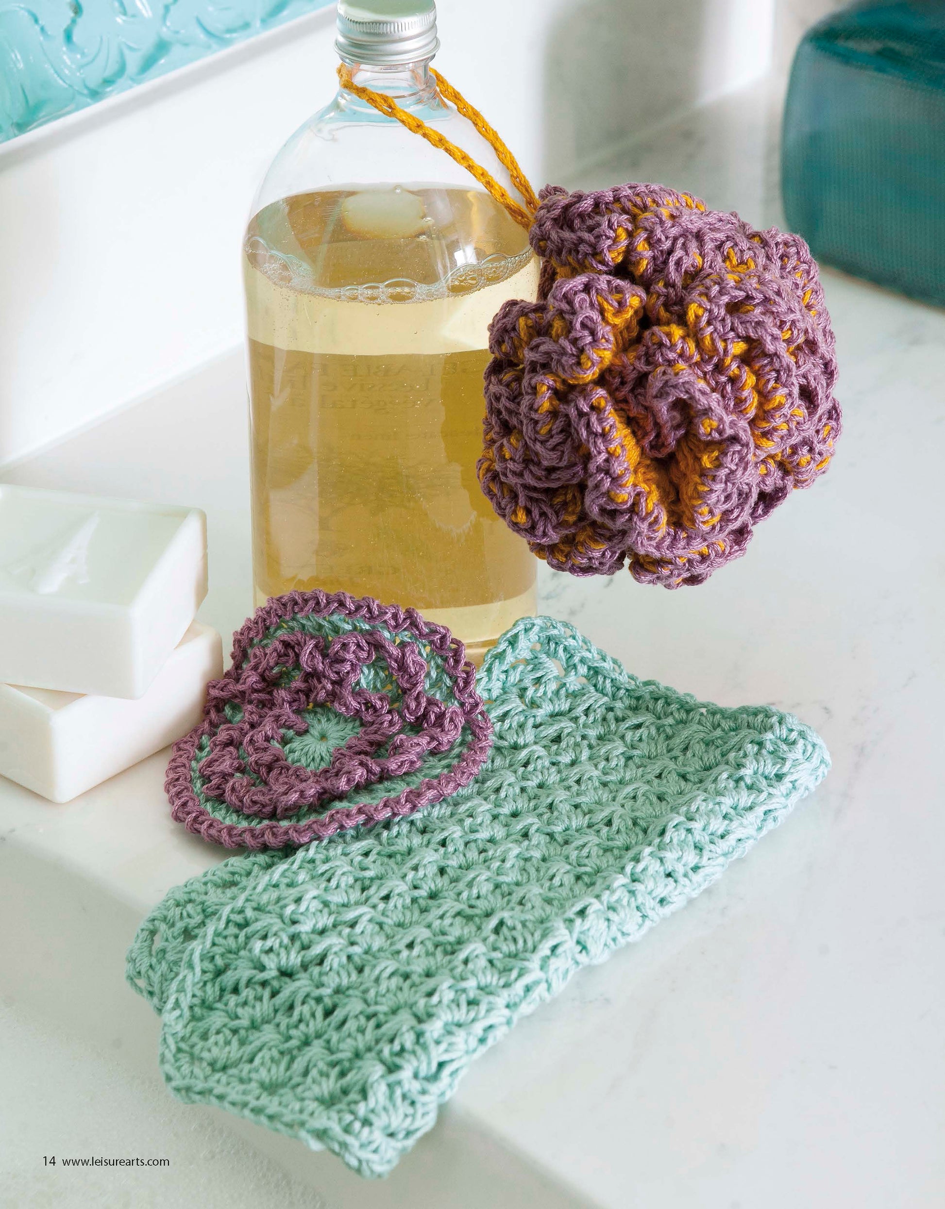 On the bathroom counter are two white soap bars, a bottle of liquid soap, a blue-green beginner washcloth, a purple and green flower, and a large purple and yellow bath pouf—all crocheted using All About Cotton by Leisure Arts.