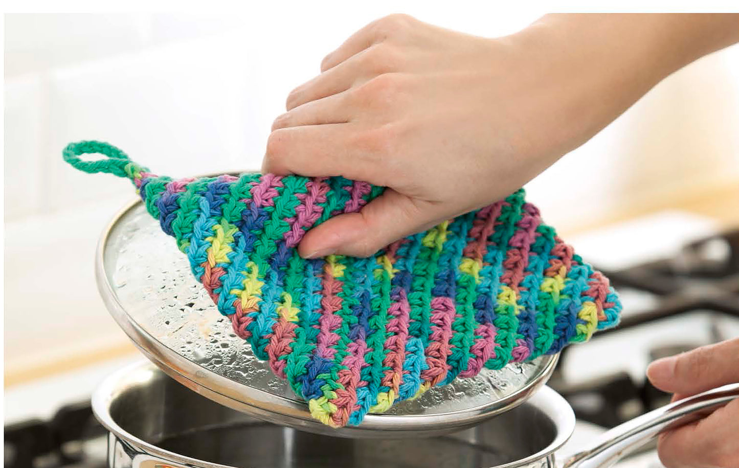 A hand uses a colorful, crocheted potholder made with Leisure Arts’ All About Cotton yarn to lift a pot lid—perfect inspiration for beginner crochet or fun projects.