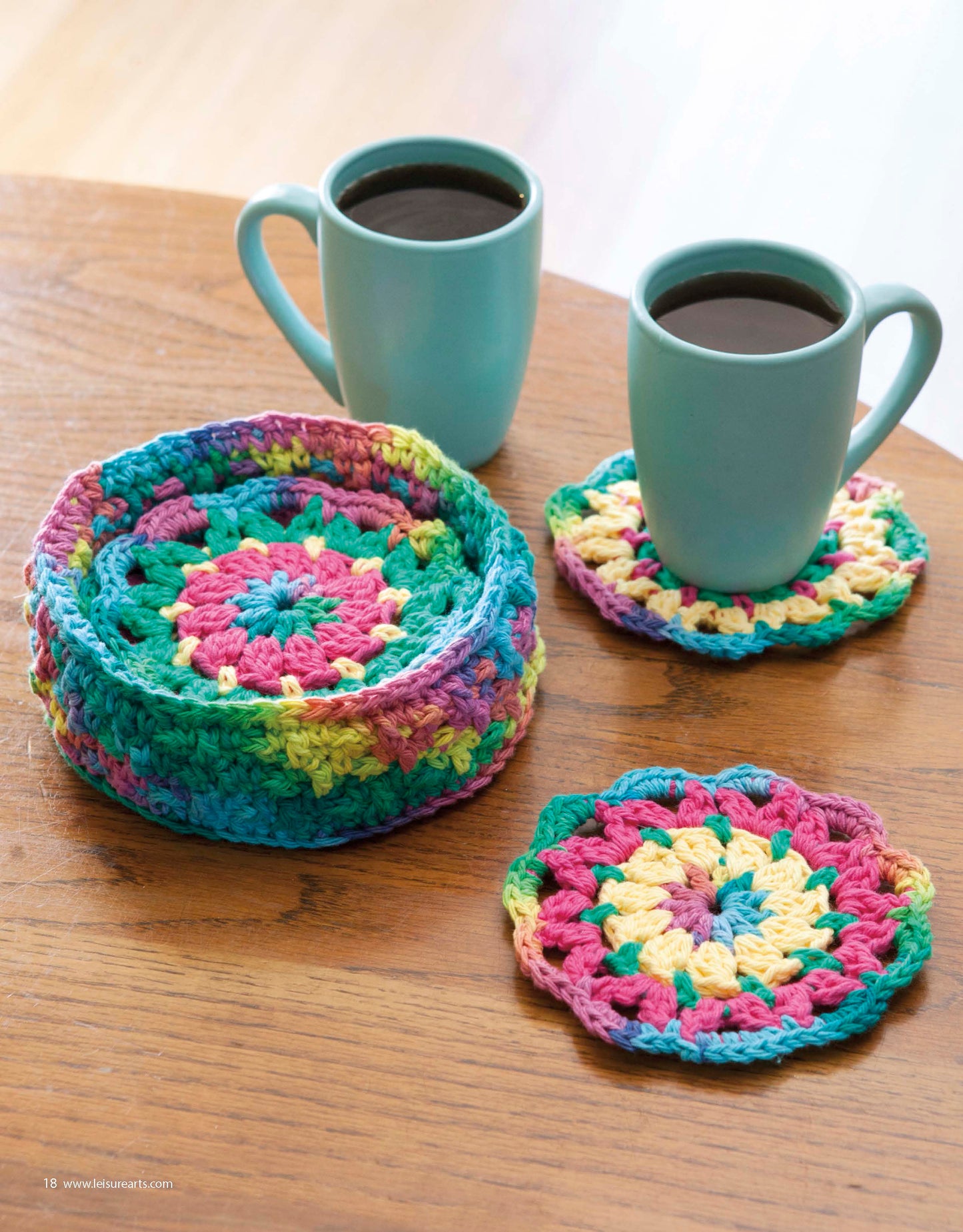 Two teal mugs of coffee sit on colorful, round coasters—beginner crochet projects made with vibrant All About Cotton yarn from Leisure Arts. A matching crocheted basket holds more coasters atop a wooden surface.