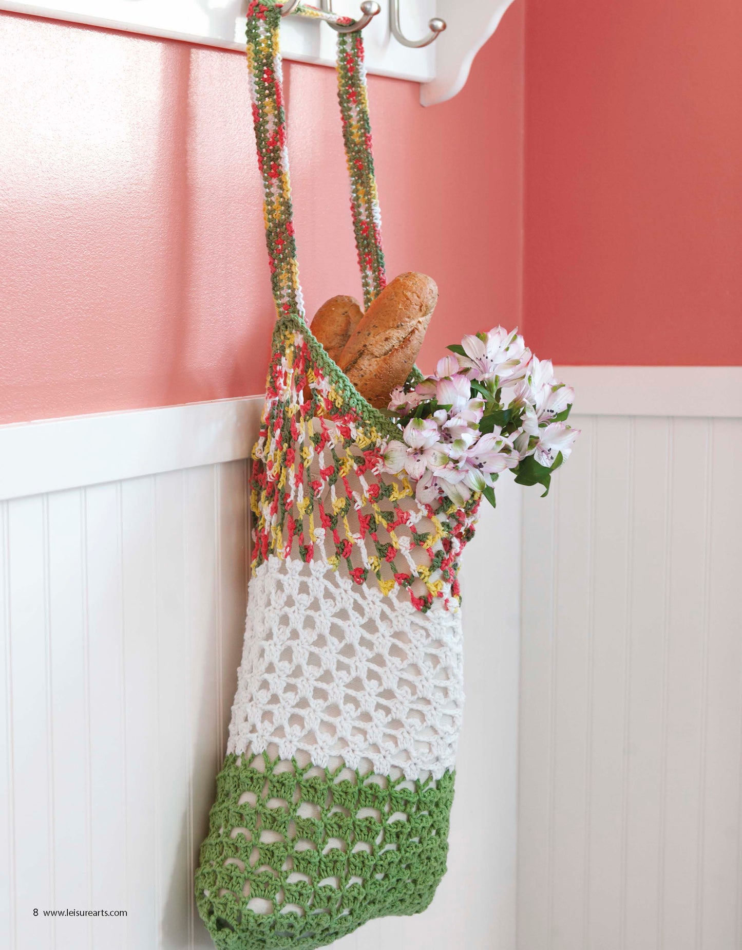 The "All About Cotton" bag by Leisure Arts, handmade from cotton yarn in green, white, and multicolor sections, hangs on a hook and holds two baguettes and pink flowers against a white and coral wall.
