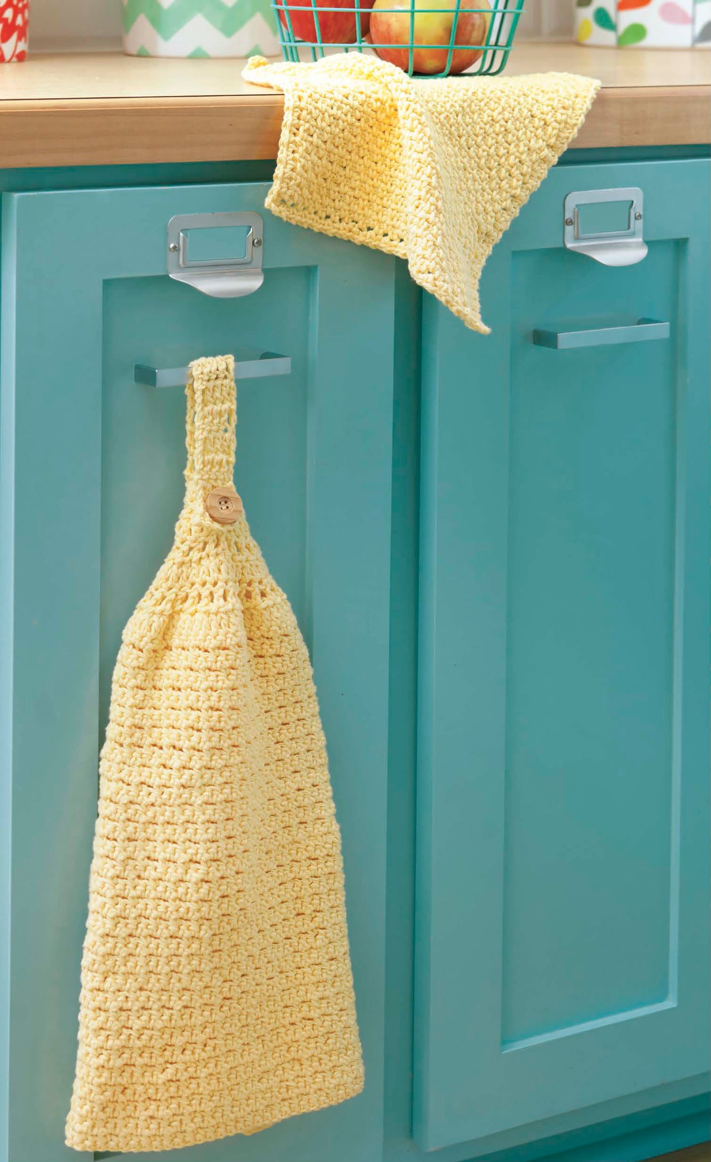 A yellow crocheted towel made with All About Cotton by Leisure Arts hangs from a button loop on a teal kitchen cabinet. A matching cloth sits on the countertop above, next to a wire basket of apples. Perfect for beginner crochet projects.