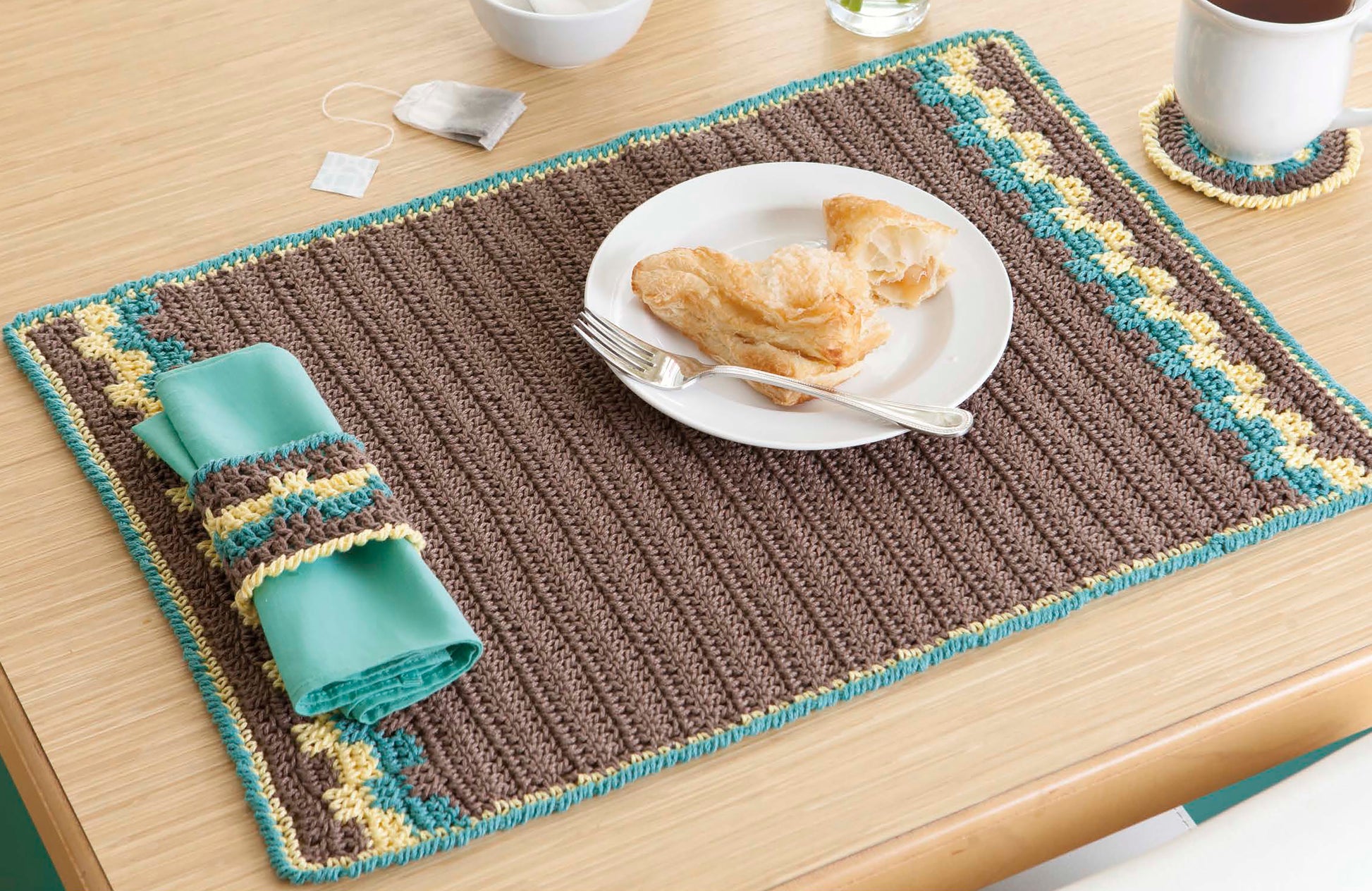 A cozy placemat made with All About Cotton by Leisure Arts in brown, yellow, and turquoise stripes holds a croissant plate, utensils, matching napkin holder, coaster with cup, and tea bag on a wooden table—perfect for beginner crochet projects.