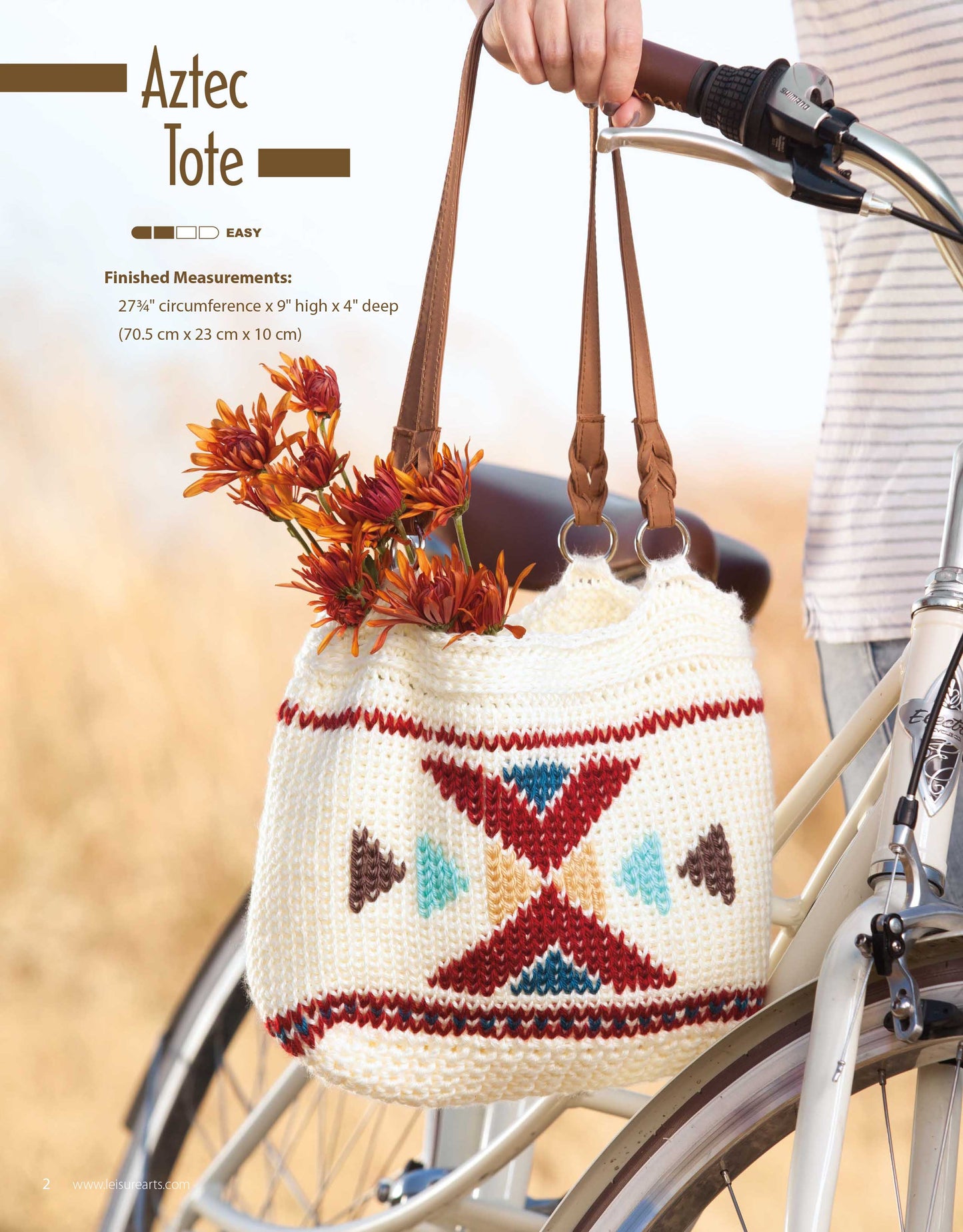 A stylish Leisure Arts Crochet Rucksacks & Backpacks design with Aztec-inspired patterns hangs from a bike handlebar by leather straps, filled with autumn flowers. Part of a hand and the white bike frame are visible. Digital download pattern.