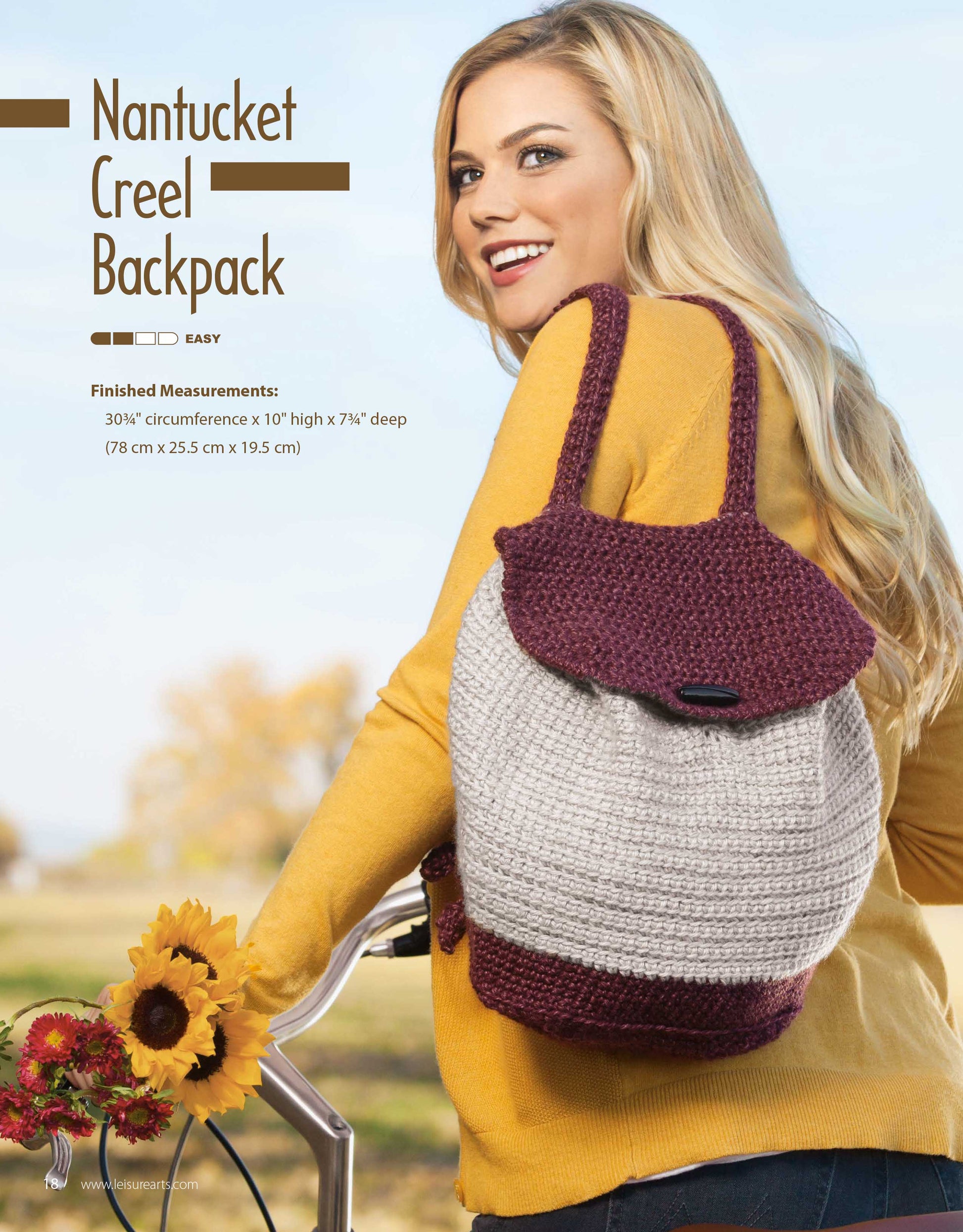 A smiling woman outdoors leans on a bicycle, holding a two-tone maroon and gray backpack. The Leisure Arts Crochet Rucksacks & Backpacks Digital Download is perfect for easy projects and stands out against autumn trees and blue sky.