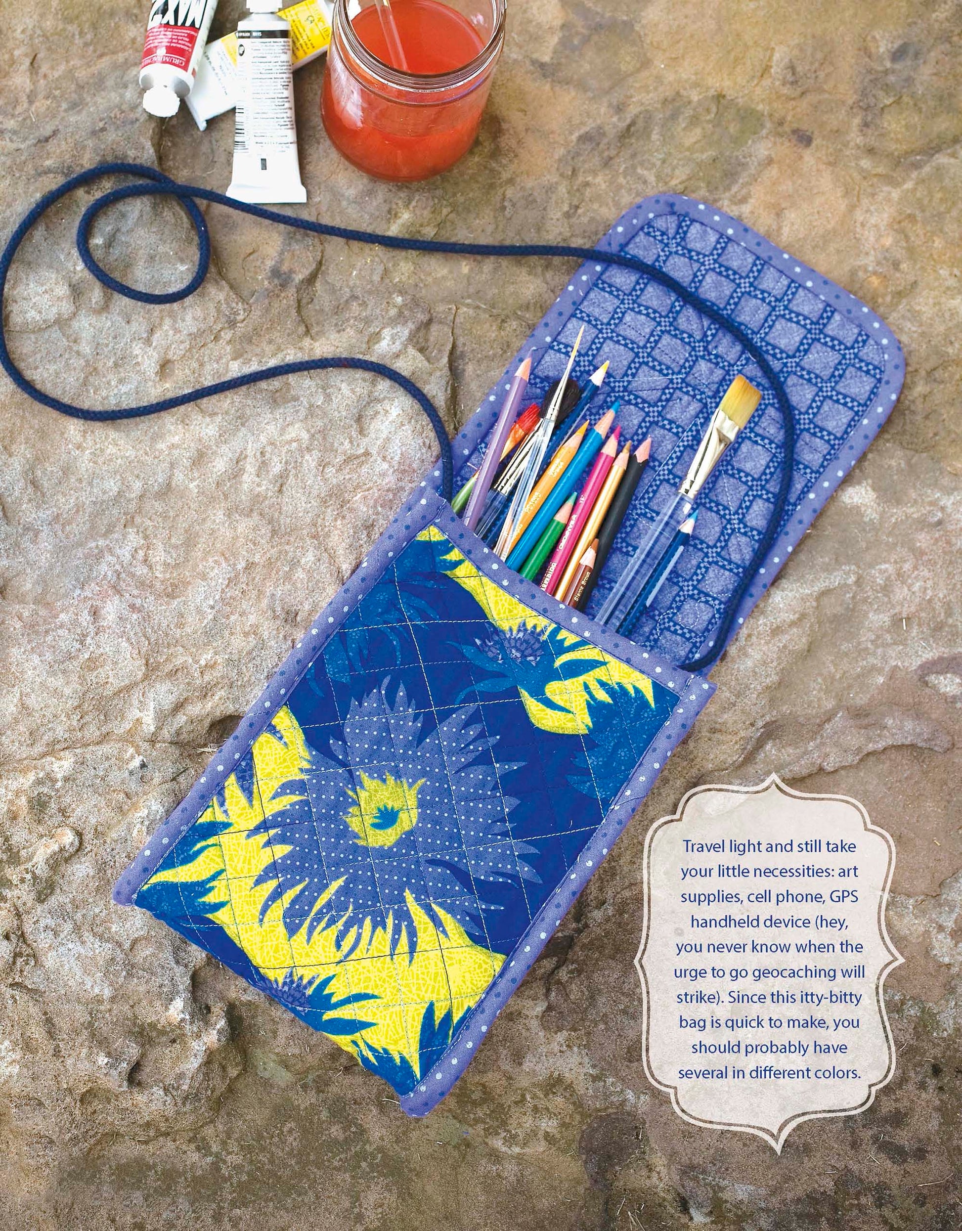 A fabric pouch made from Leisure Arts' Best of Quilted Bags Digital Download rests open on stone, holding colored pencils and a paintbrush. Nearby are tubes of paint and a jar with pink liquid and a paintbrush inside.