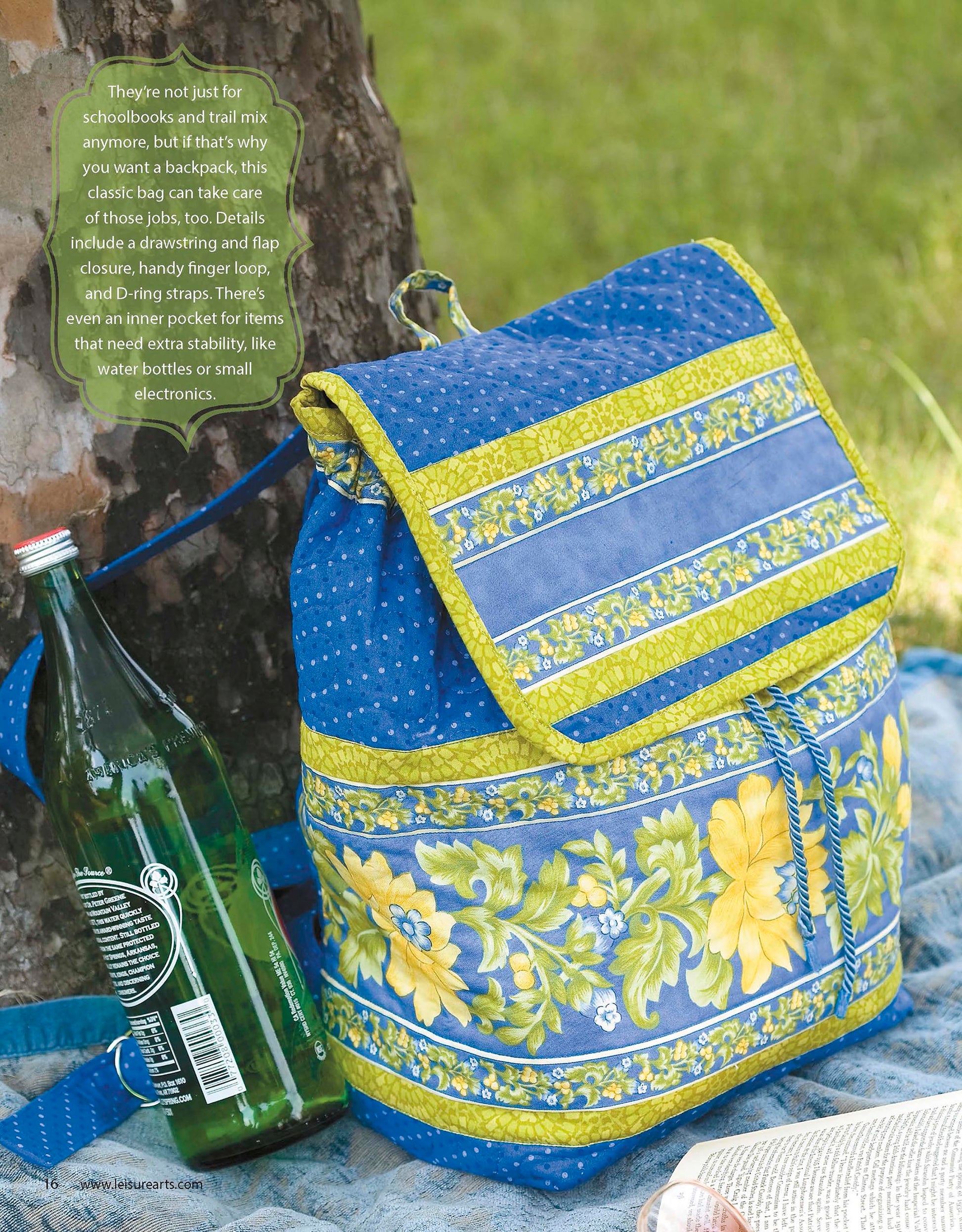 A vibrant floral fabric backpack rests on a blue blanket with a glass bottle and tree trunk—perfect inspiration for crafting your own look with the Leisure Arts Best of Quilted Bags Digital Download.