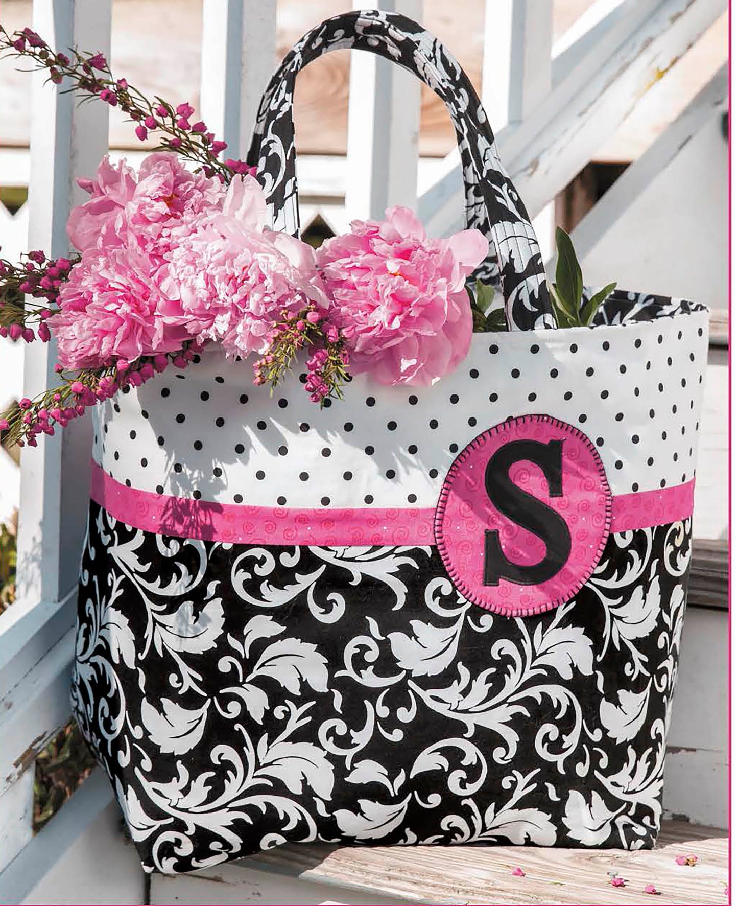 The Best of Quilted Bags Digital Download by Leisure Arts features a stylish black and white tote with polka dot accents, a pink ribbon, and an S patch filled with pink peonies—ideal for beginners seeking creative quilted bag patterns.